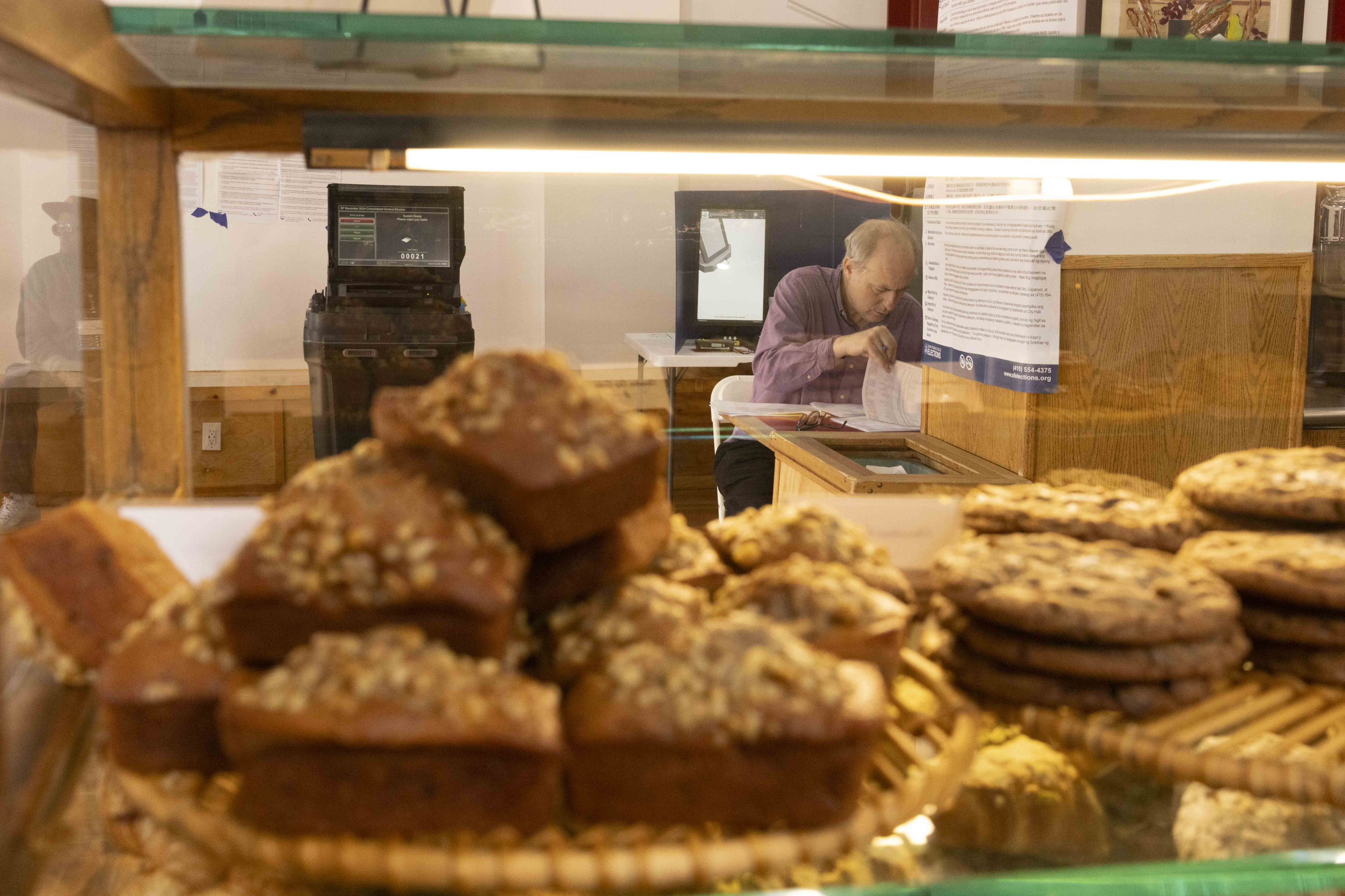 A man is sitting behind a glass display filled with muffins and cookies. He appears to be handling paperwork, with a voting machine visible in the background.