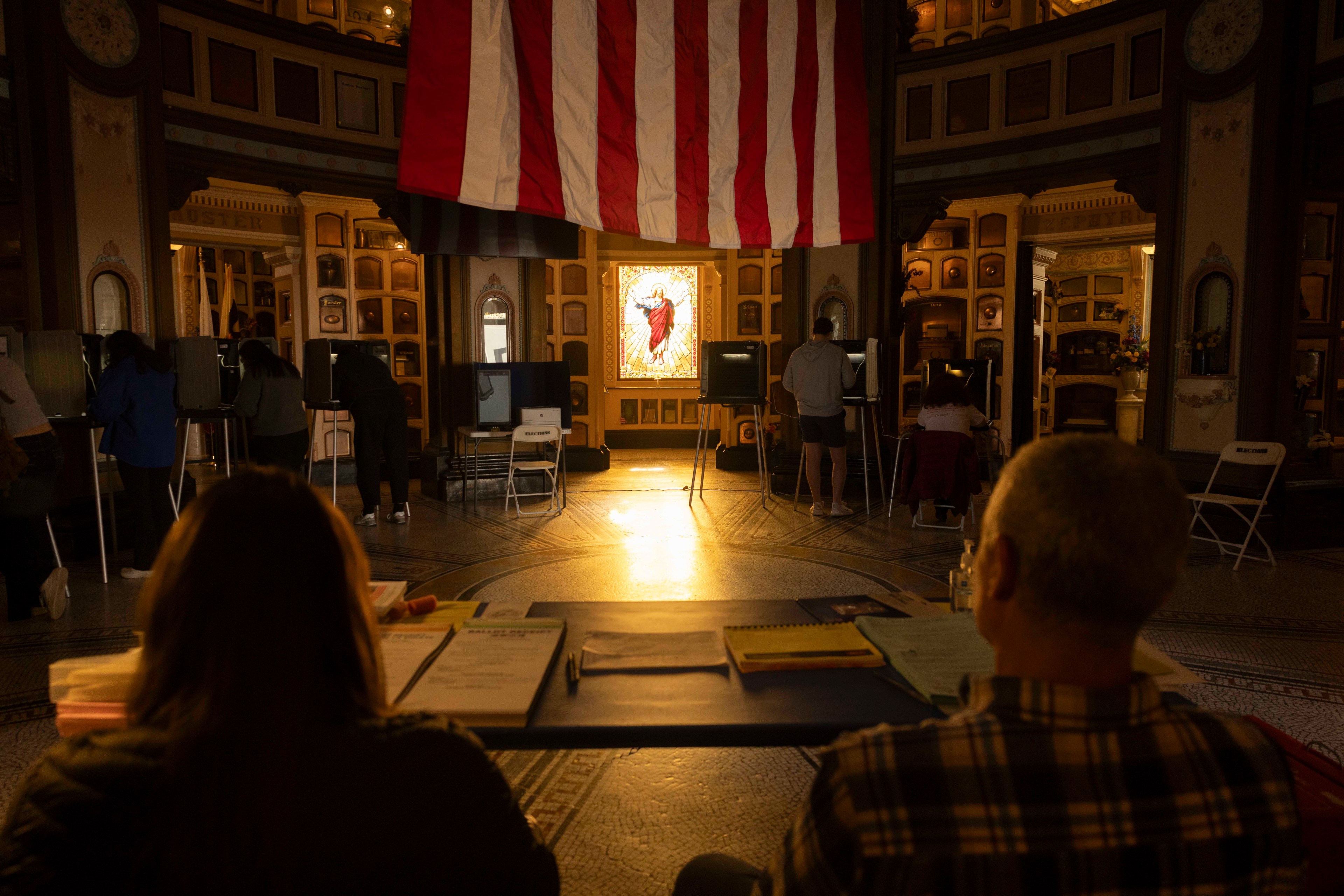 People are voting at booths in a dimly lit, ornate room with a large American flag hanging and a stained glass window in the background.