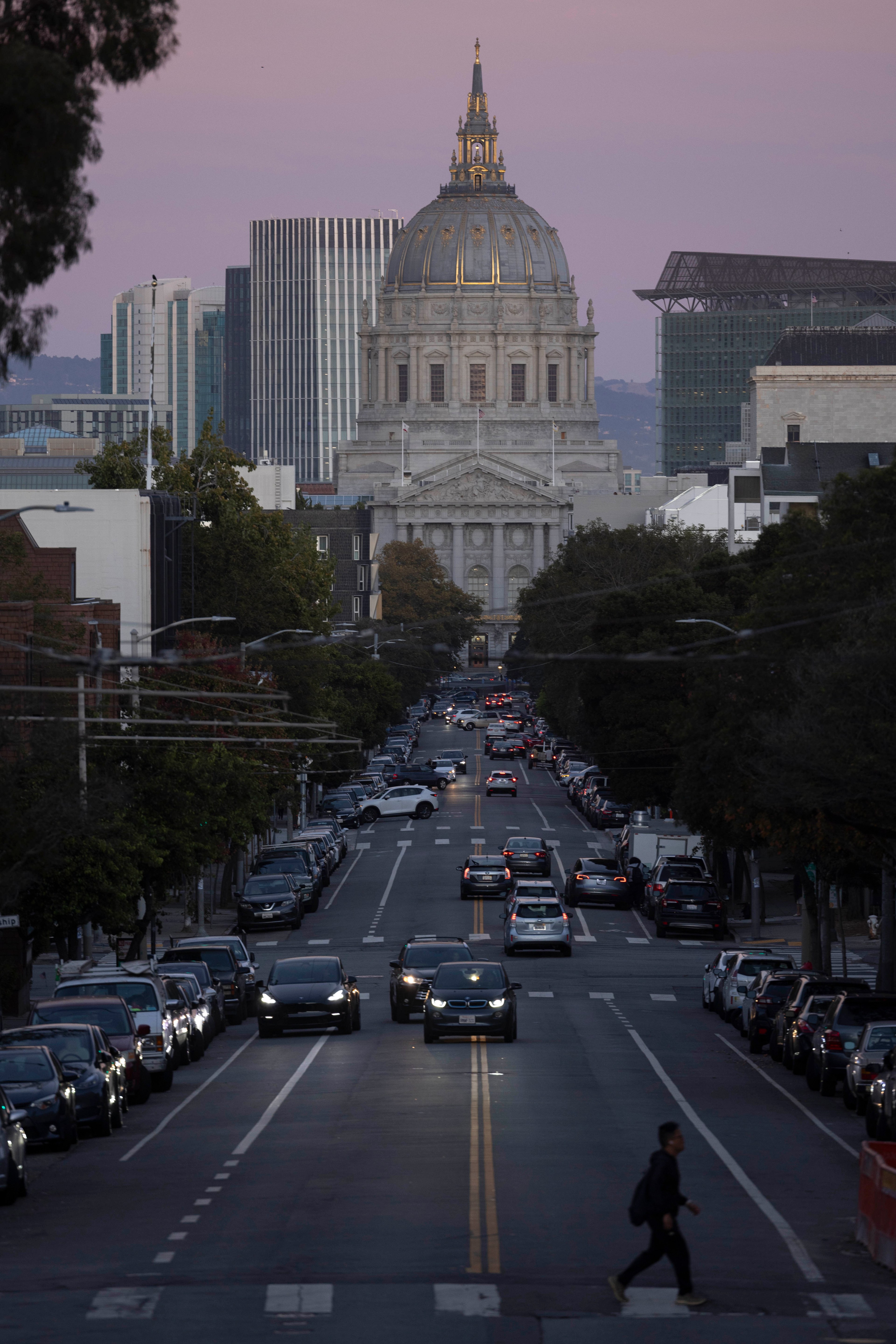 A street leads to a grand building with a dome, set against a twilight sky. Cars line the street, and a person crosses in the foreground.