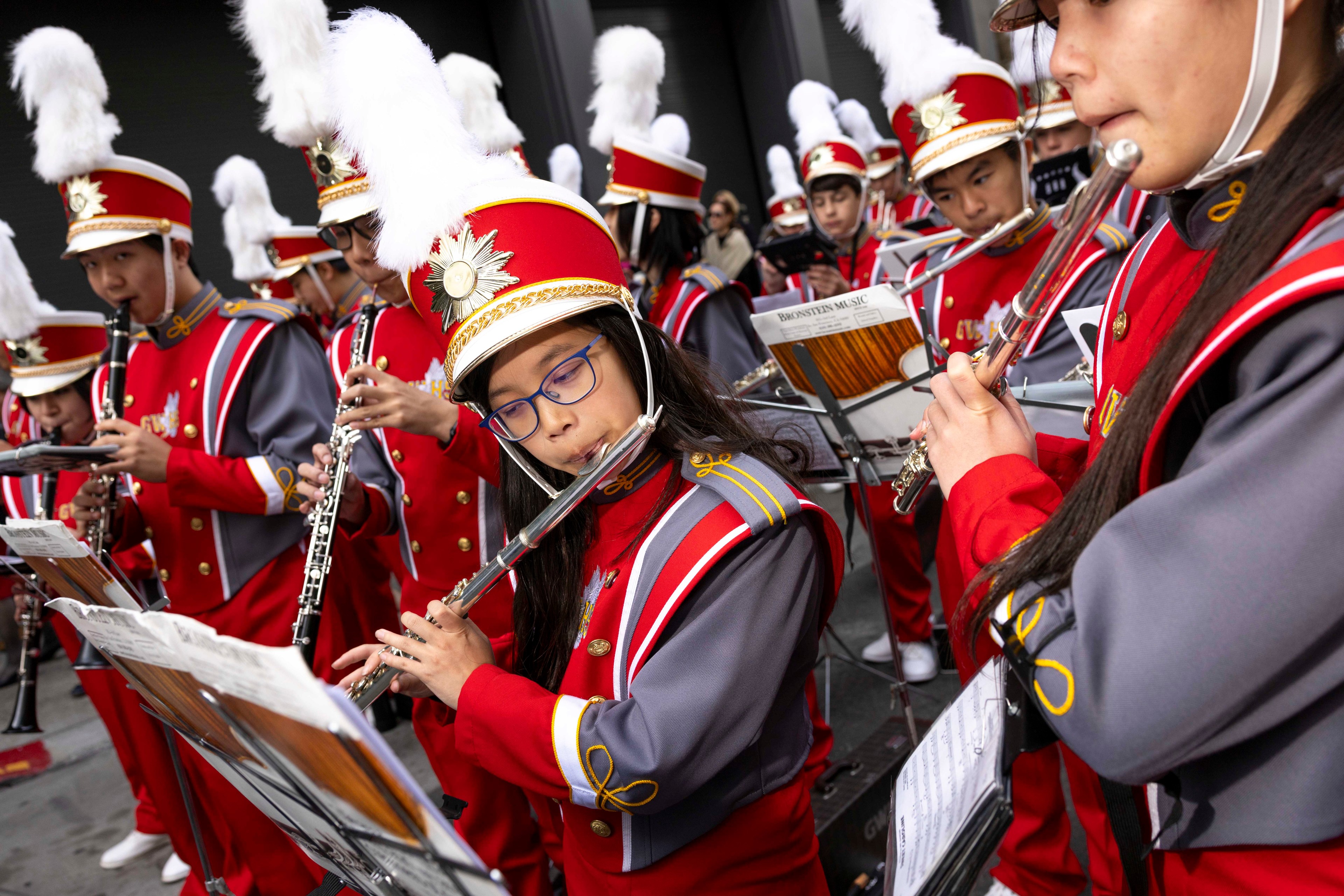 A marching band in vibrant red and gray uniforms plays instruments like flutes and clarinets, with members wearing red hats adorned with white feathers.