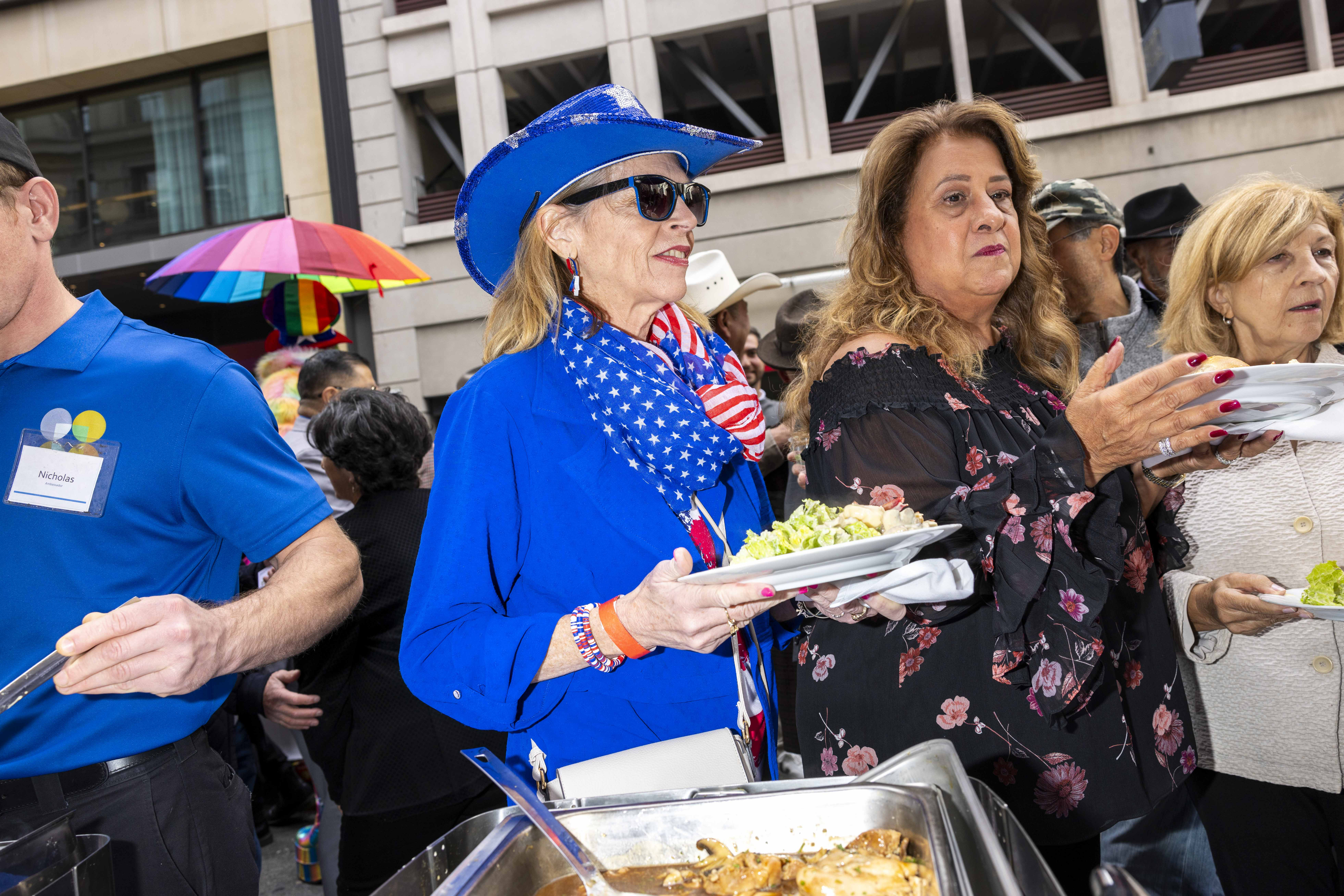 A woman in a blue outfit with a star-spangled scarf and cowboy hat holds a plate of food. She stands next to a man serving and a crowd under a colorful umbrella.