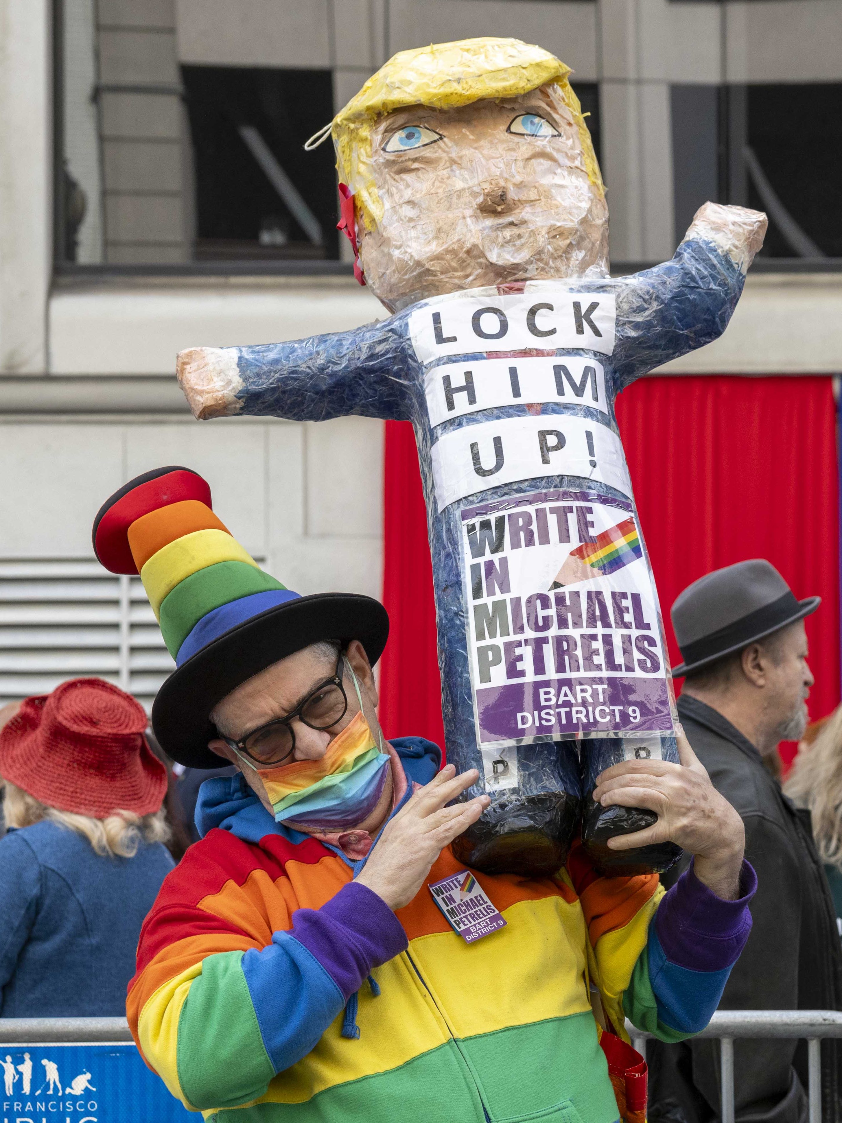 A person in a rainbow outfit holds a large puppet with yellow hair. The puppet has a &quot;LOCK HIM UP!&quot; sign, and the person wears a rainbow mask and hat.