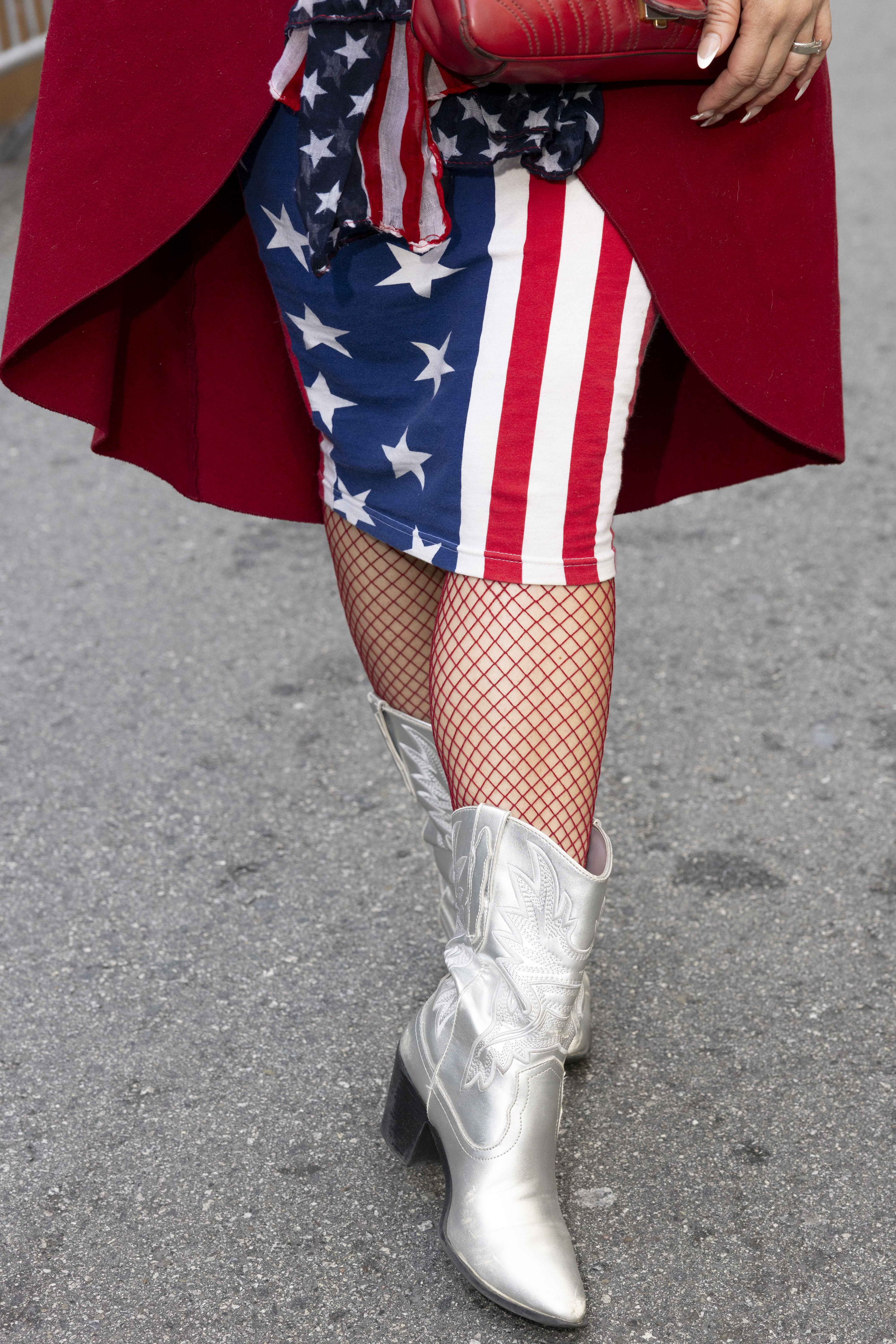 A person wears a red coat, a skirt with the American flag design, red fishnet stockings, silver cowboy boots, and holds a red purse against an asphalt background.