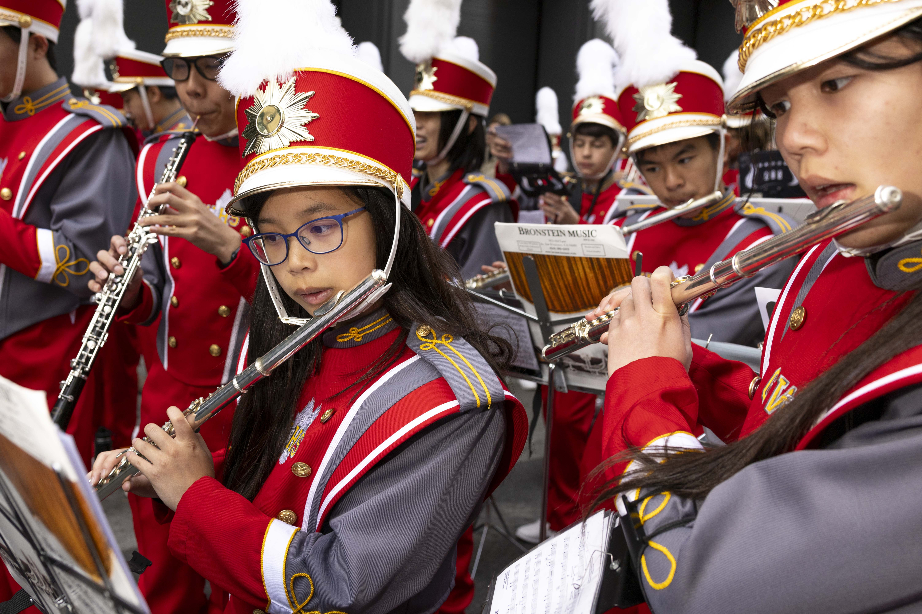 A marching band in red and gray uniforms, with feathered hats, plays flutes and other instruments while reading sheet music.