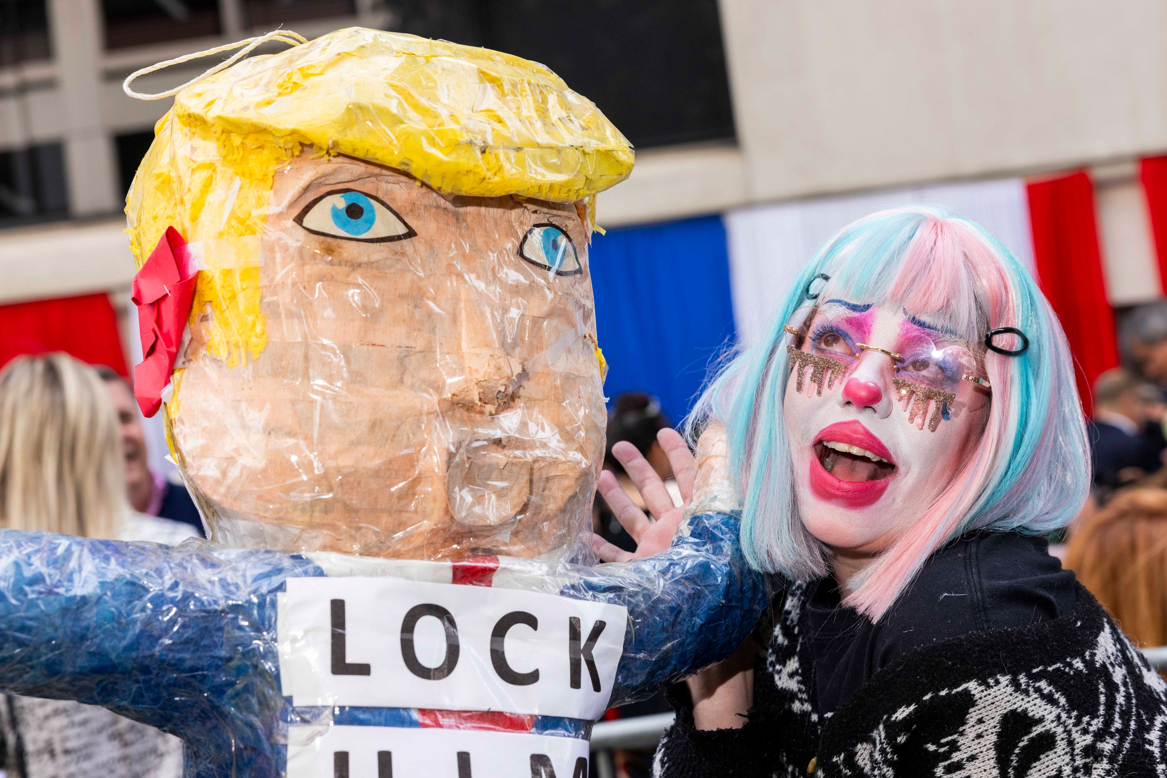 A colorful clown with a pink and blue wig and white face paint poses next to a large paper-mâché figure wearing a sign with the text "LOCK HIM UP."