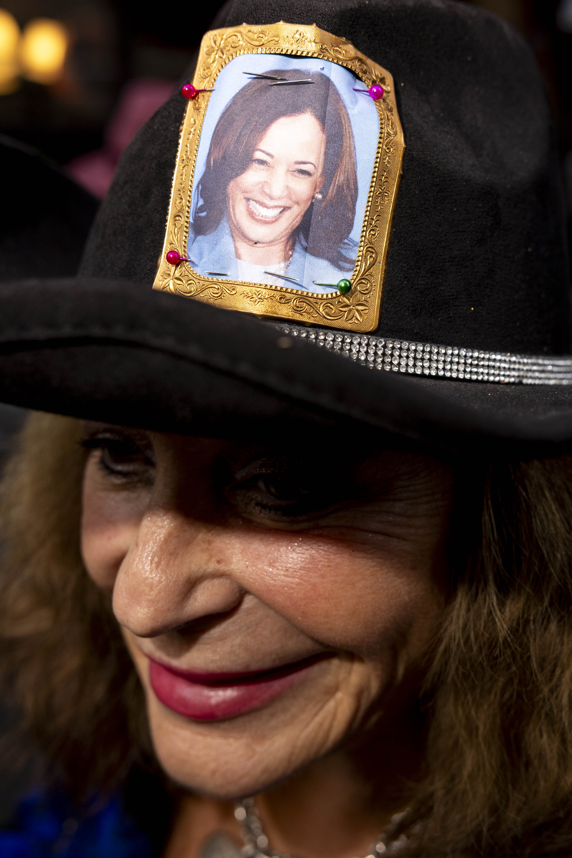 A woman wears a black hat adorned with a jeweled band and a pinned photo of another woman smiling. Brightly colored pins secure the photo.