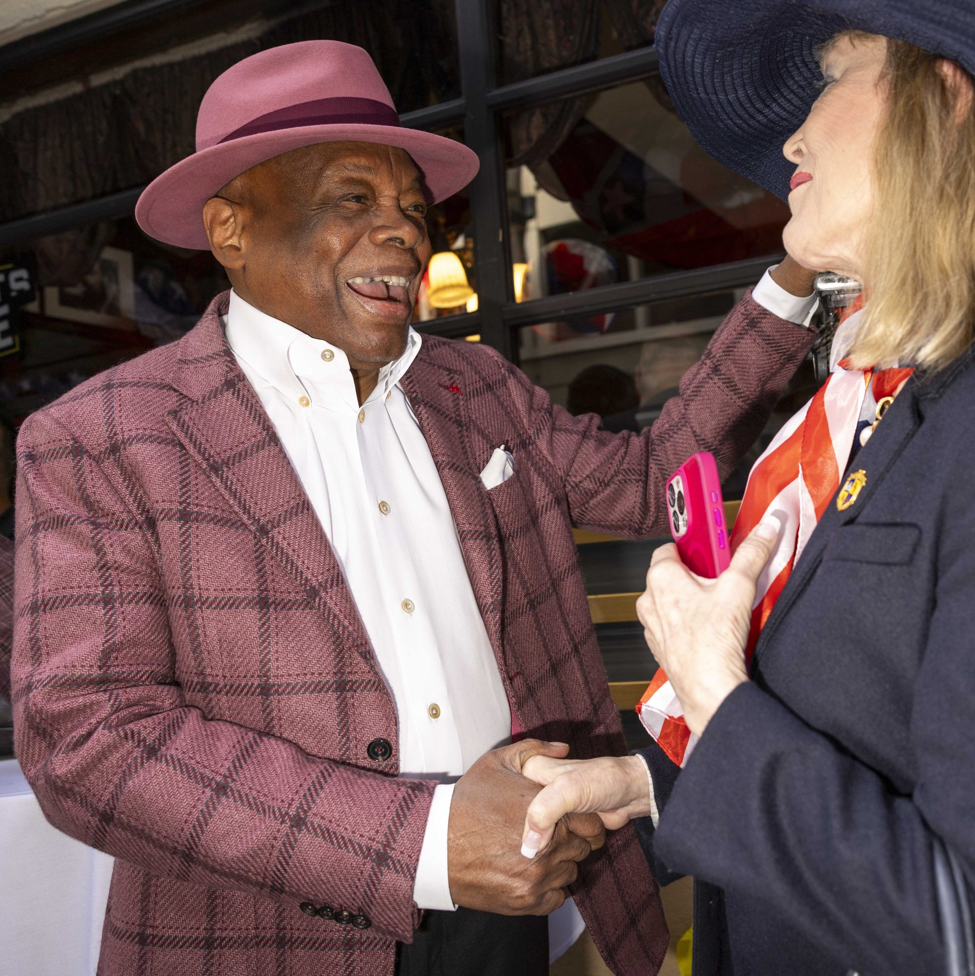 A man wearing a pink hat and plaid jacket is joyfully shaking hands with a woman in a navy outfit holding a pink phone, both standing indoors.