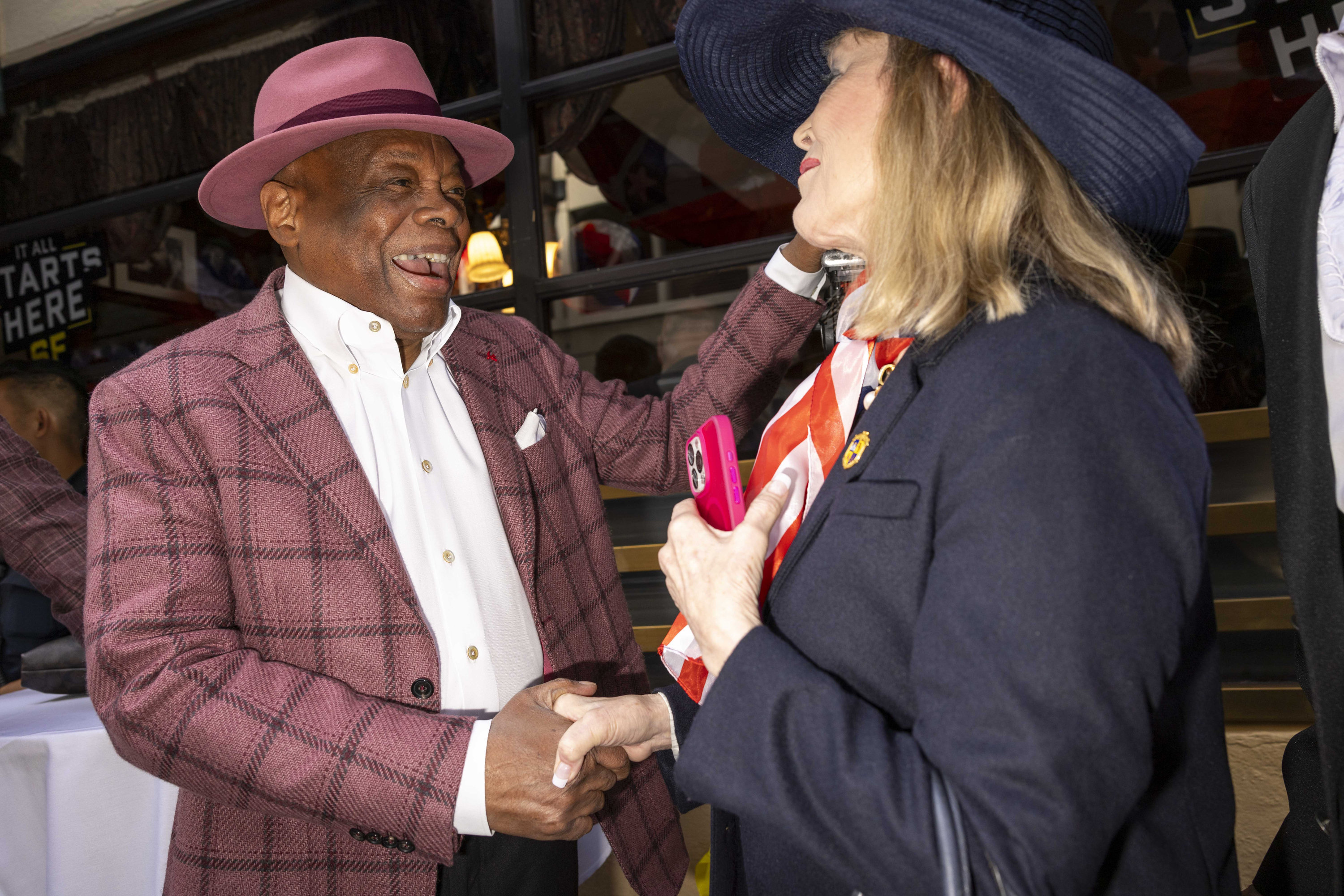 Two people are warmly greeting each other, both wearing stylish hats. The man in a pink hat and checked jacket is smiling broadly, shaking hands with the woman in a blue hat.