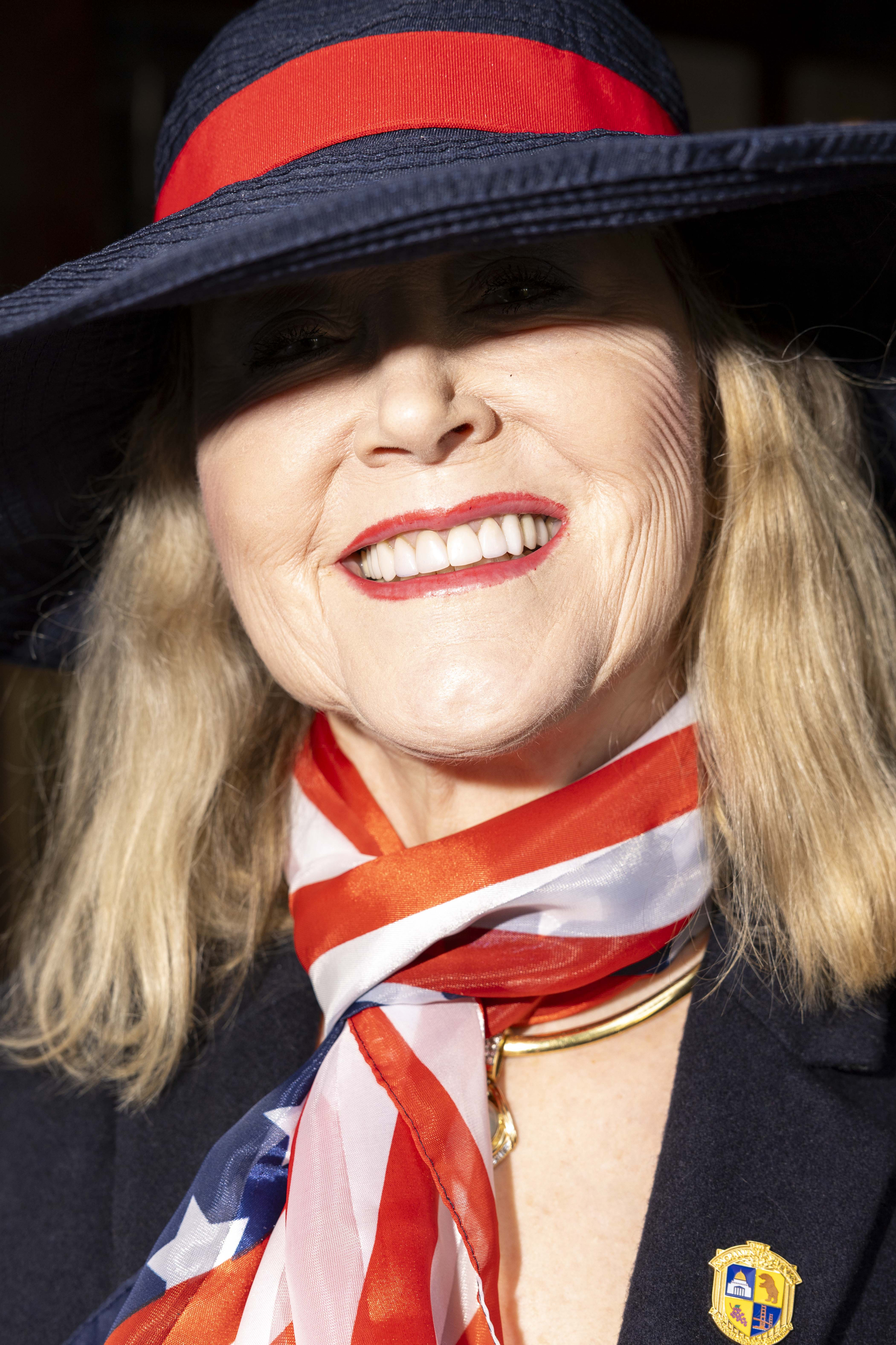 A person is smiling and wearing a blue hat with a red band, a scarf with an American flag pattern, and a dark coat with a gold pin.