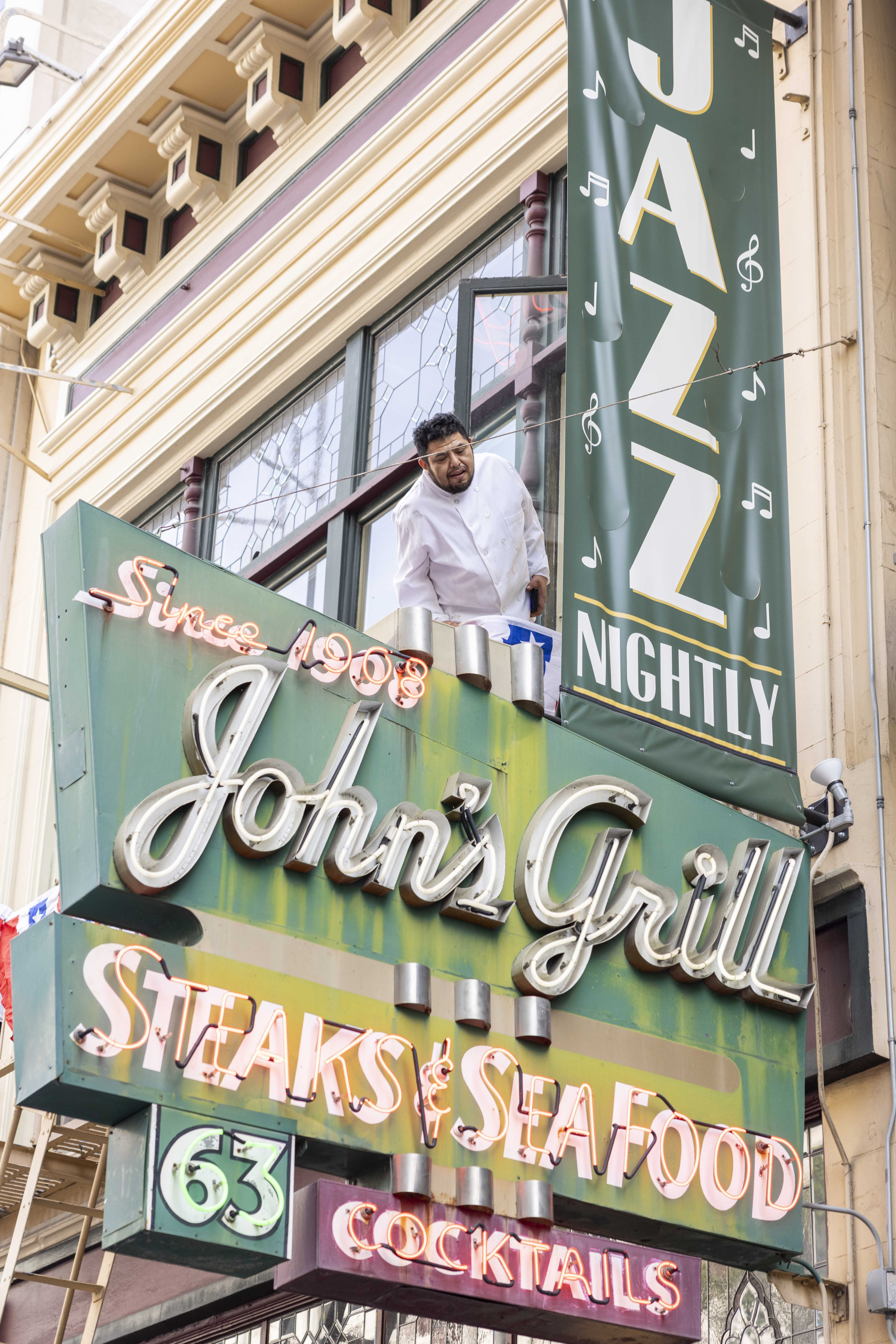 A man leans out of a window above a vintage neon sign reading &quot;John's Grill,&quot; with &quot;Steaks &amp; Seafood&quot; and &quot;Cocktails.&quot; A banner says &quot;Jazz Nightly.&quot;