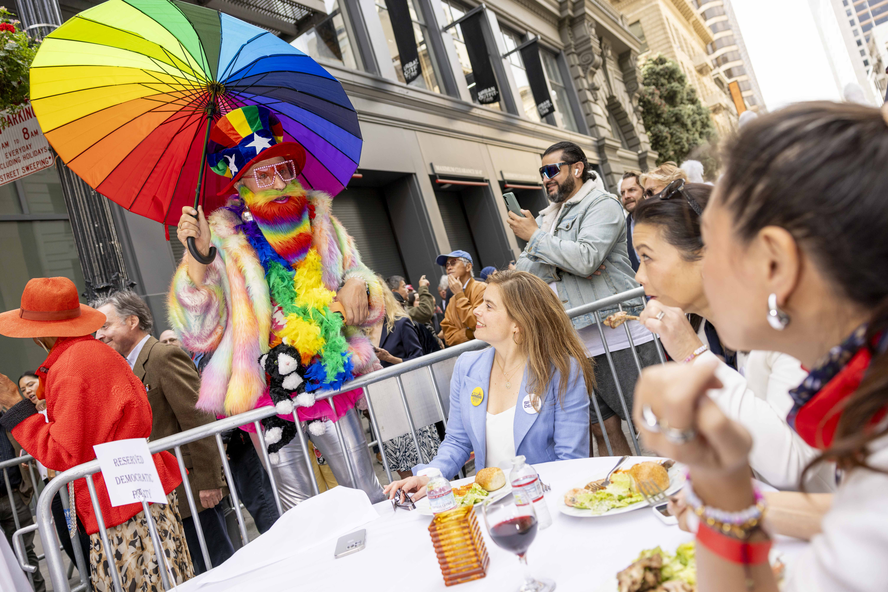 A person in a rainbow outfit and hat holds a colorful umbrella, engaging with seated diners near a street barrier. People around are taking photos and observing.