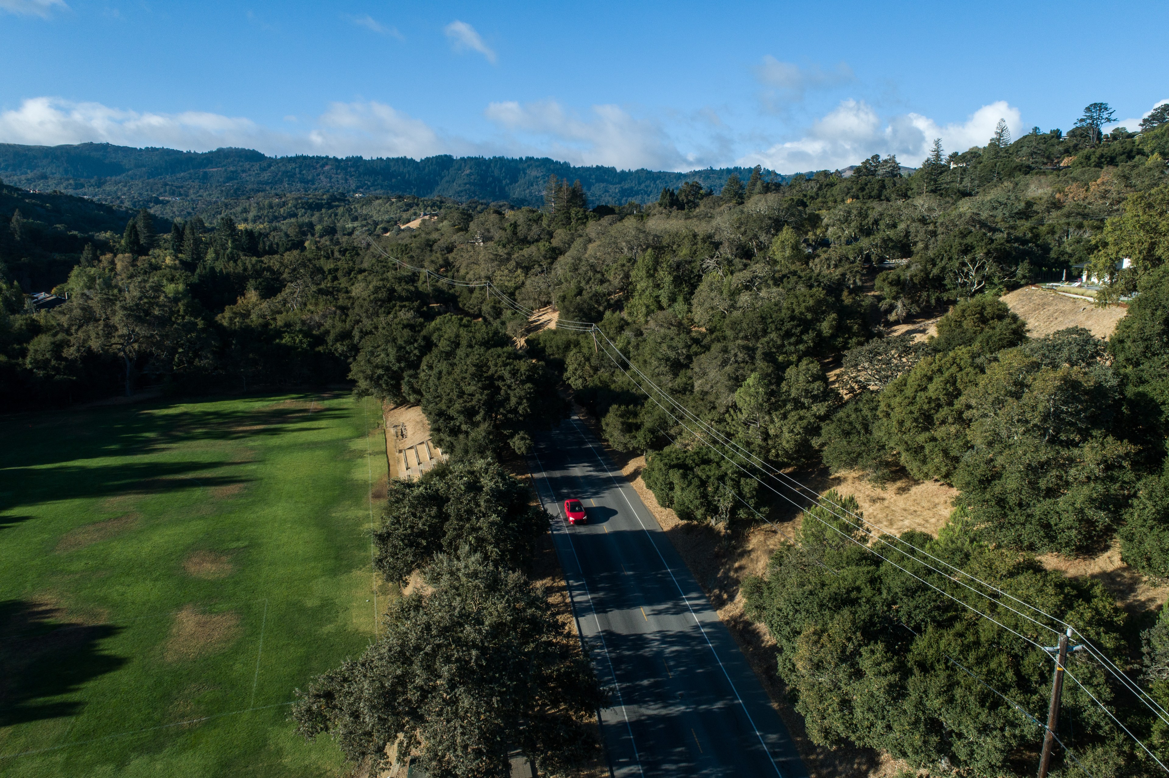 A red car drives along a road flanked by dense green trees. To the left, there’s a lush green field. Hills and mountains are visible under a clear blue sky.