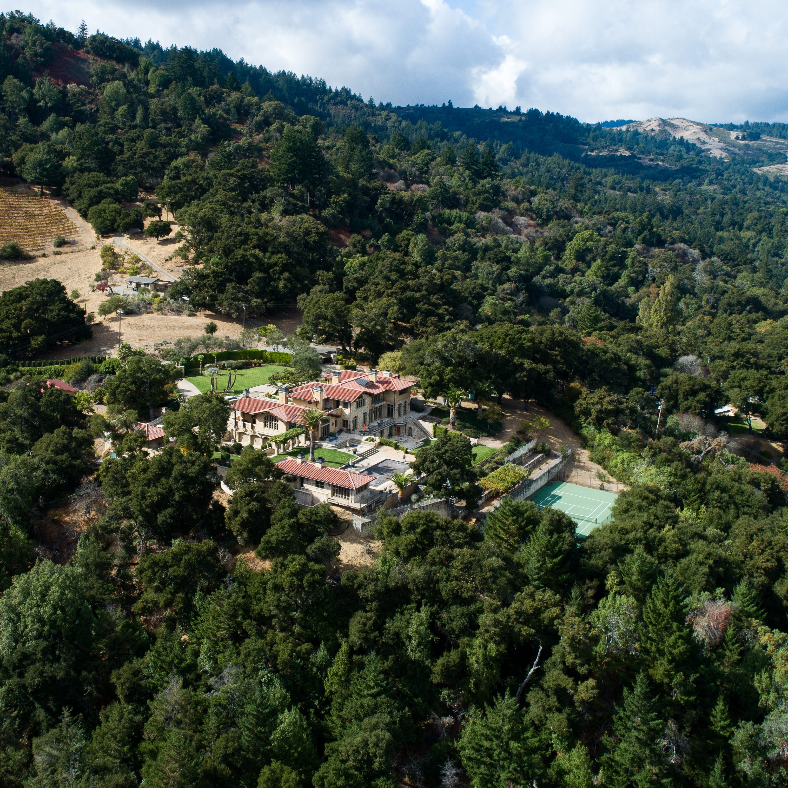 A large estate is nestled among lush trees, featuring a grand house with a red-tiled roof, manicured lawns, a swimming pool, and a tennis court.