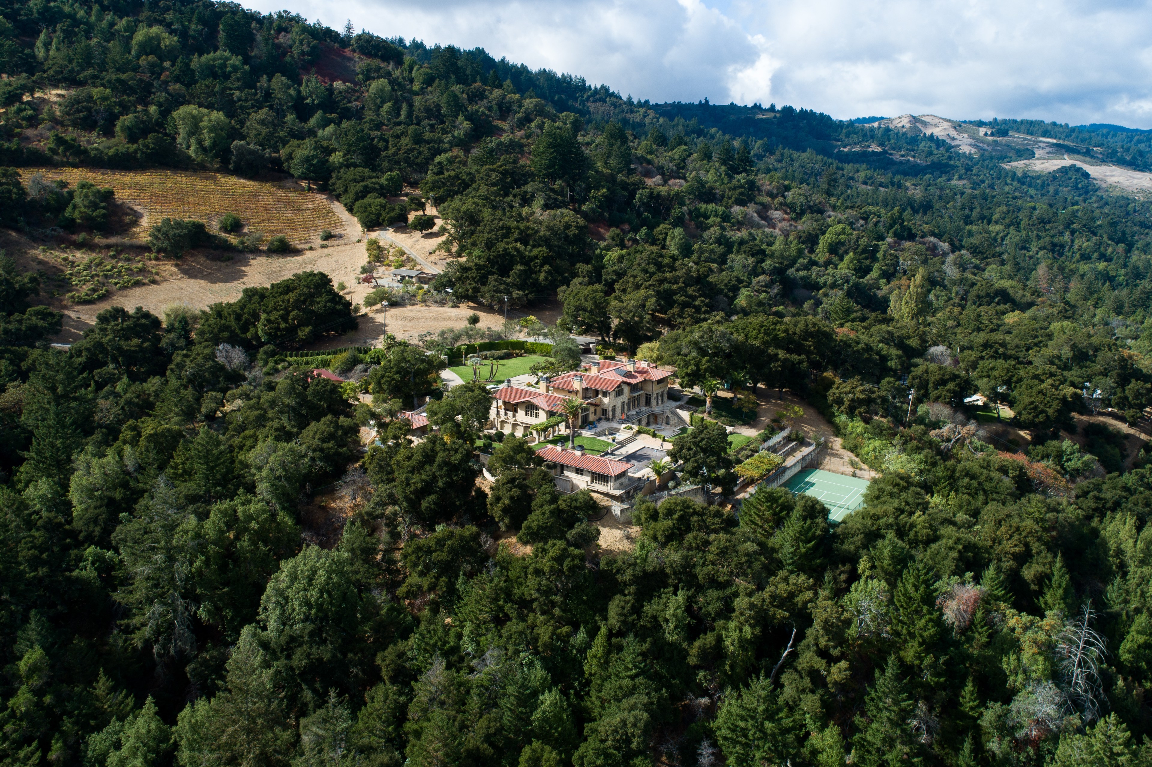 The image shows a large villa with a red-tiled roof nestled among dense, rolling green hills. A tennis court is visible nearby, surrounded by lush trees.