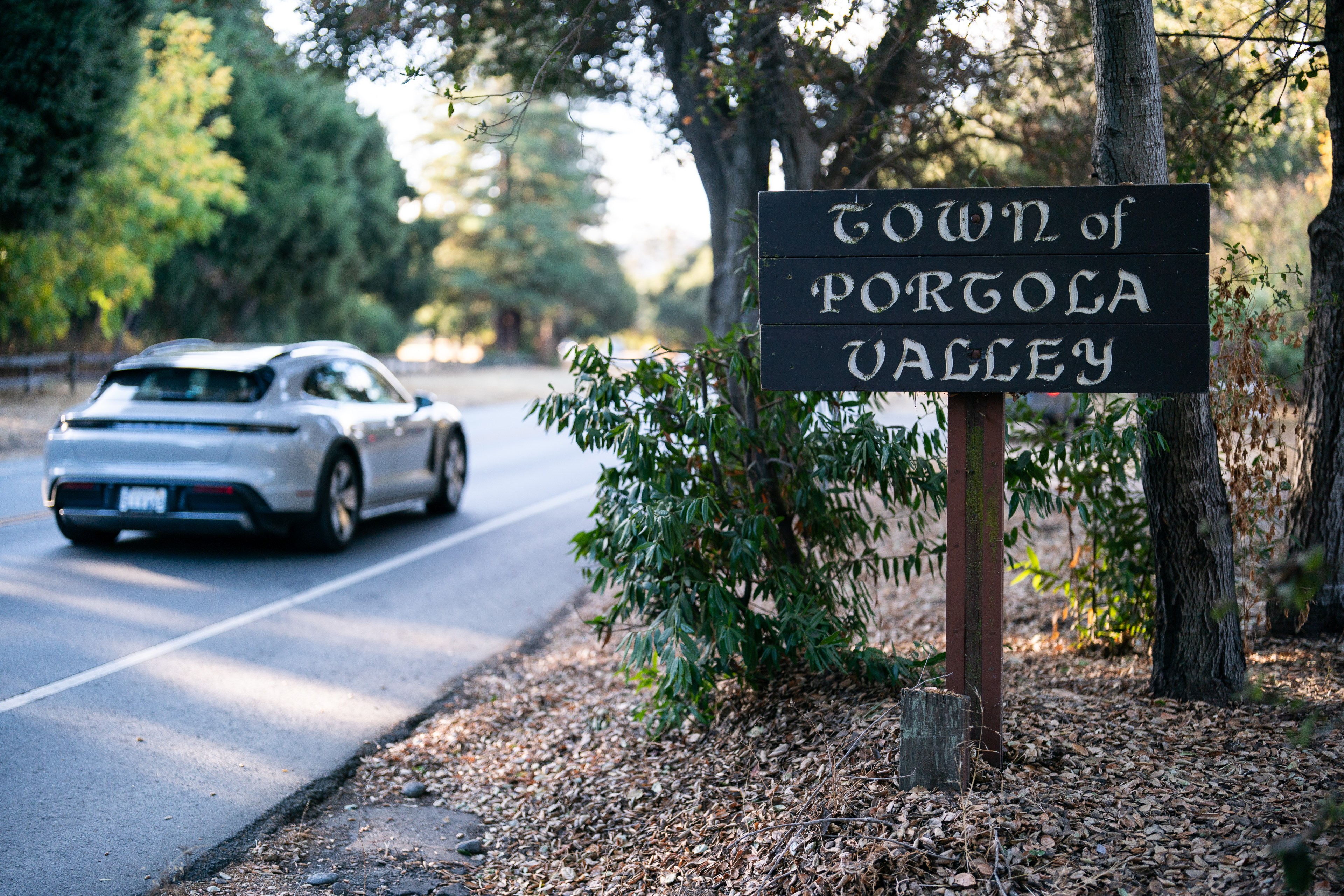 A silver car drives on a tree-lined road beside a wooden sign reading &quot;Town of Portola Valley,&quot; surrounded by greenery and fallen leaves.