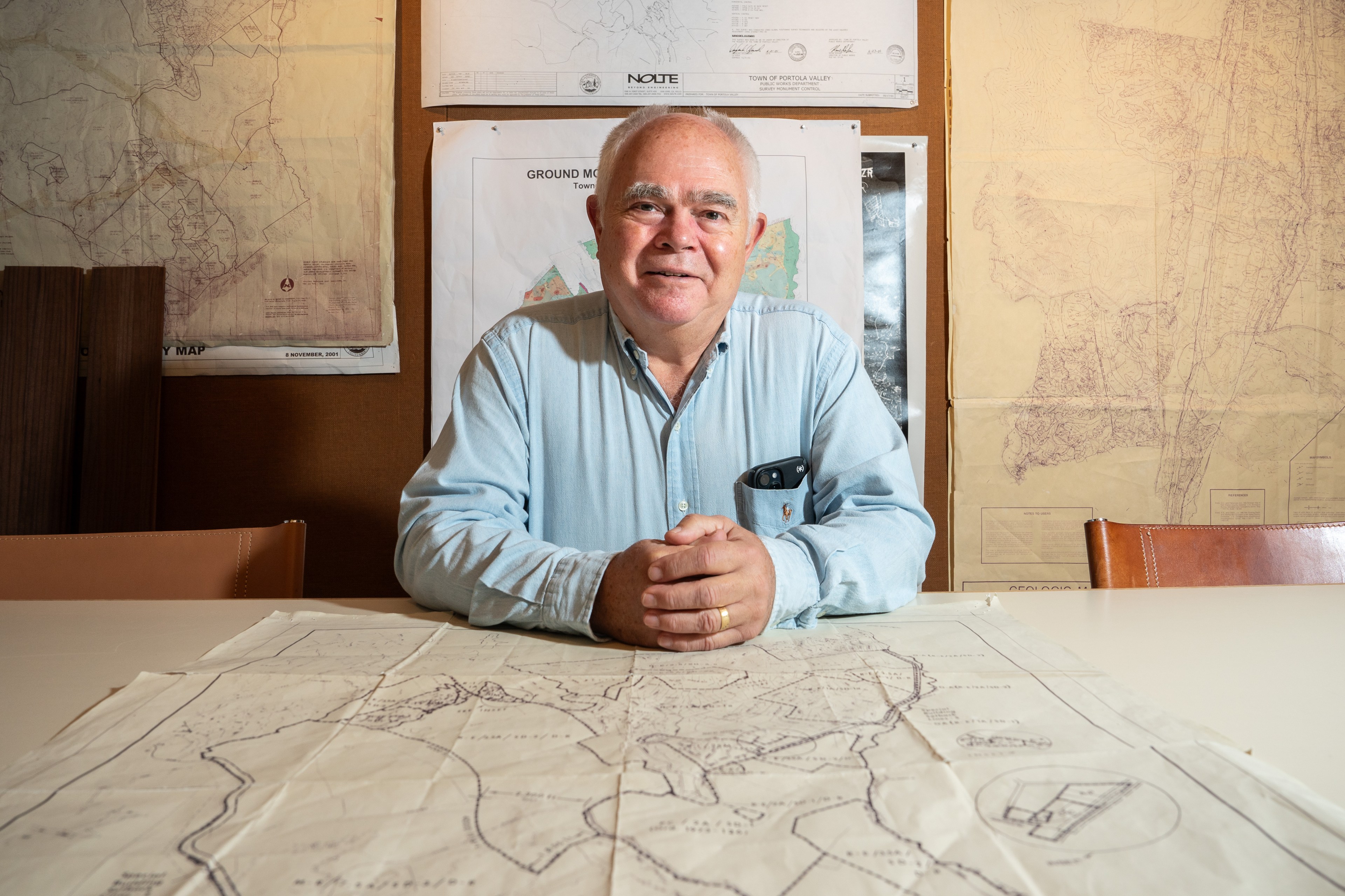 A man in a light blue shirt sits at a table covered with maps. Behind him, more maps are pinned on the wall, creating an office-like setting.