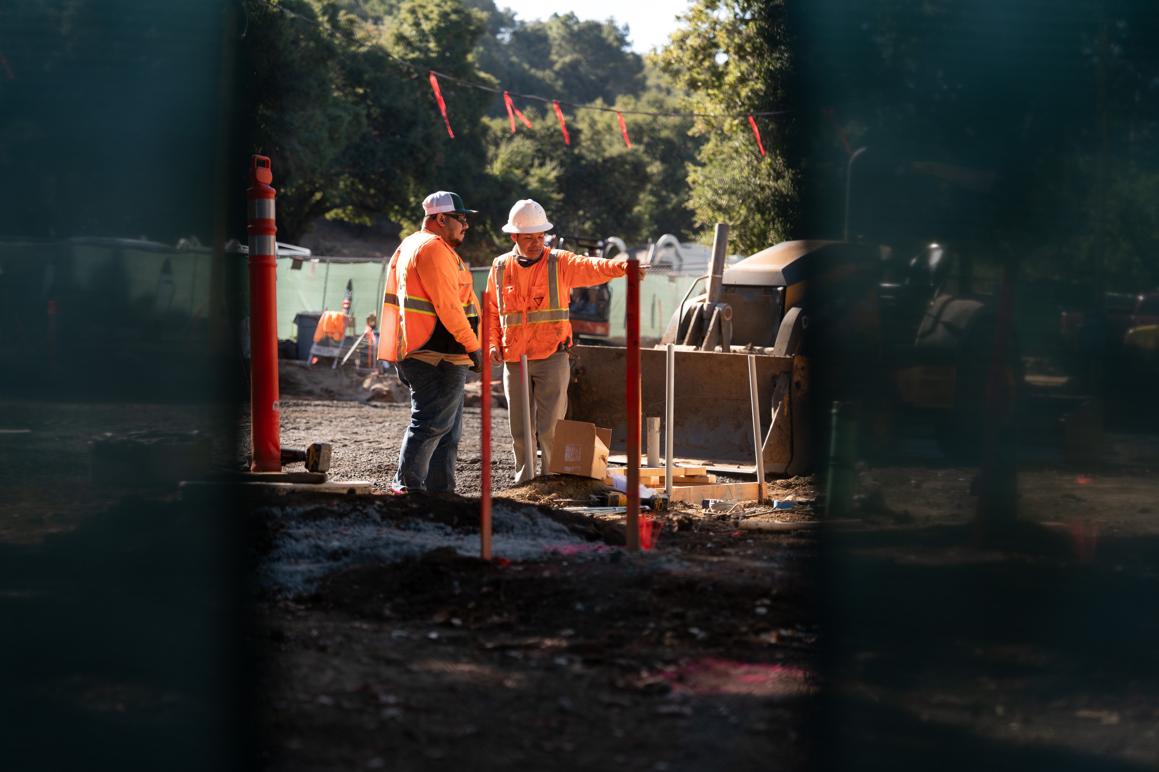 Two construction workers in orange safety vests and helmets are discussing a project at a worksite with equipment and trees in the background.