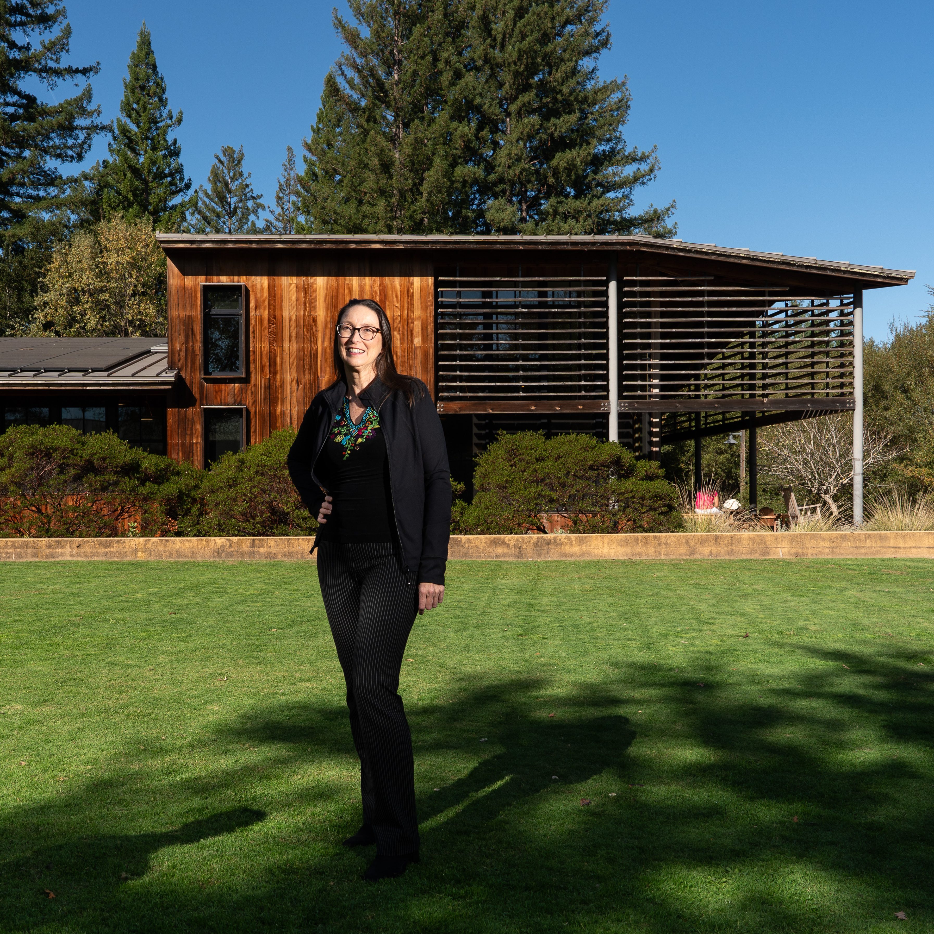 A person stands on a green lawn in front of a modern wooden house surrounded by lush greenery. They are wearing black attire and glasses, smiling at the camera.