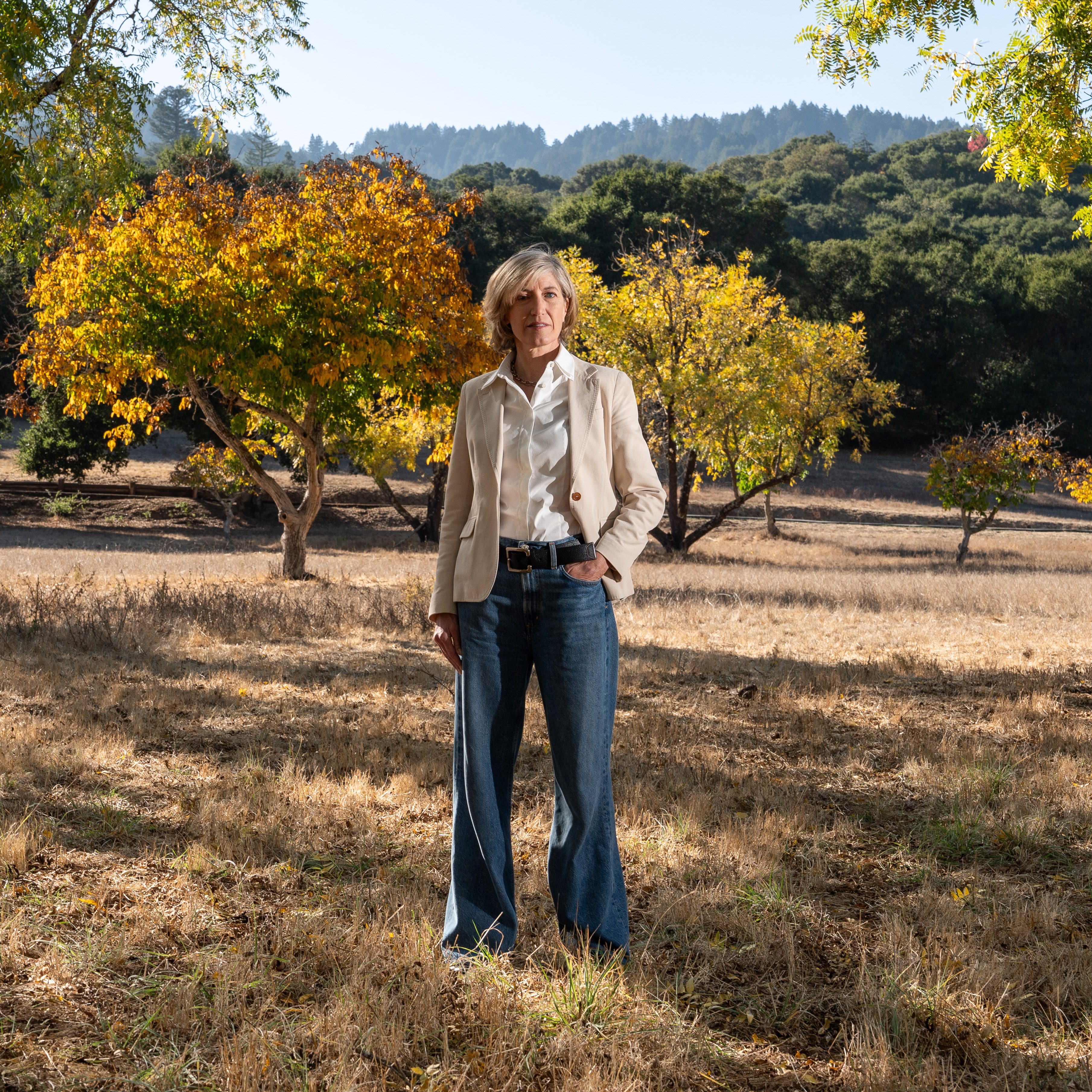 A woman stands in a dry field with fall-colored trees behind her, dressed in a beige jacket and jeans. The scene is bright, with a forested hill in the background.