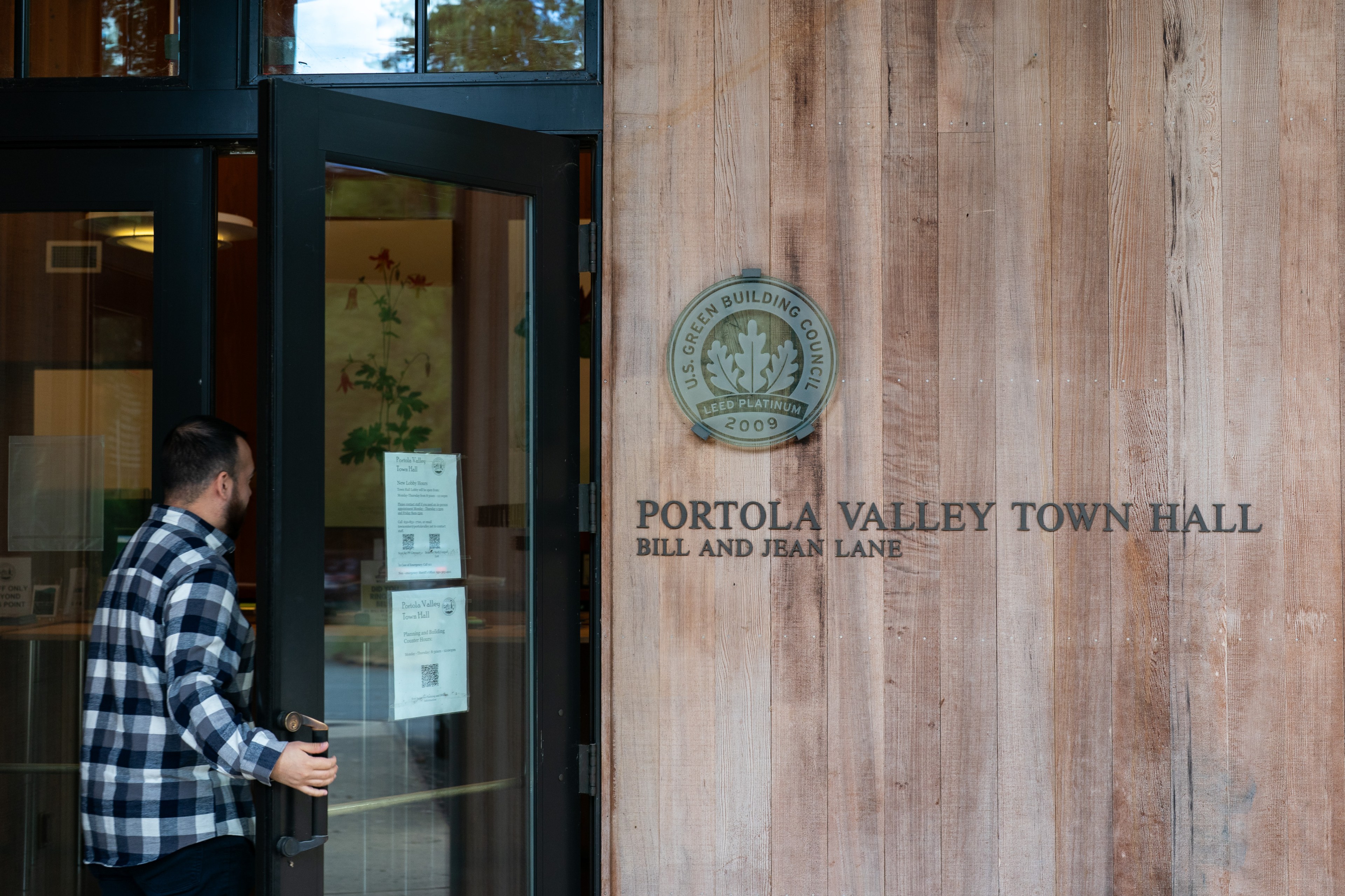 A person in a checkered shirt opens a glass door at the Portola Valley Town Hall, with a LEED Platinum 2009 plaque on the wooden wall beside it.