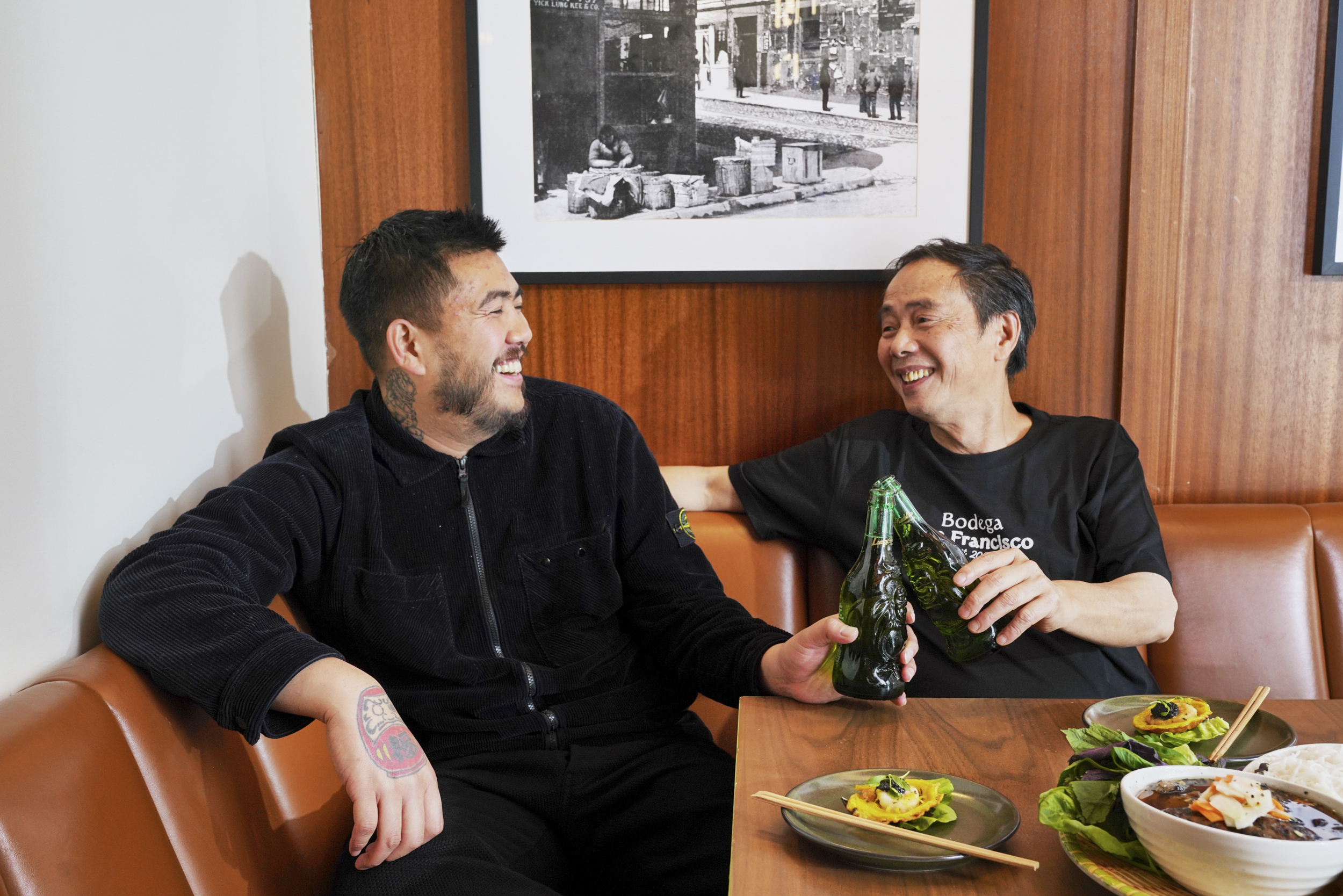 Two people are sitting on a couch, smiling and clinking green bottles. They are surrounded by plates of food and a framed black-and-white photo is on the wall behind them.