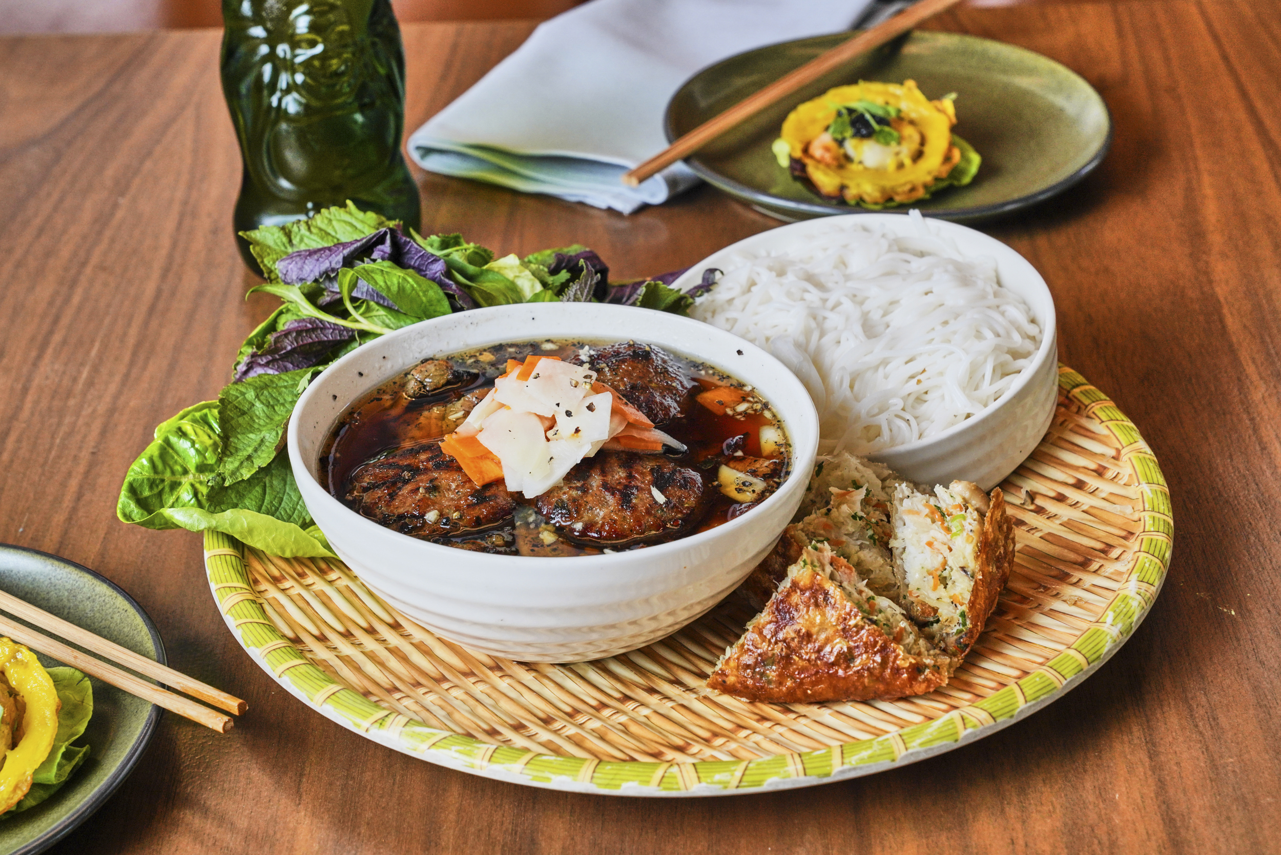 A platter displays a bowl of broth with meat patties topped with pickled veggies, rice noodles, fresh greens, and a fried savory pastry, set on a wood table.