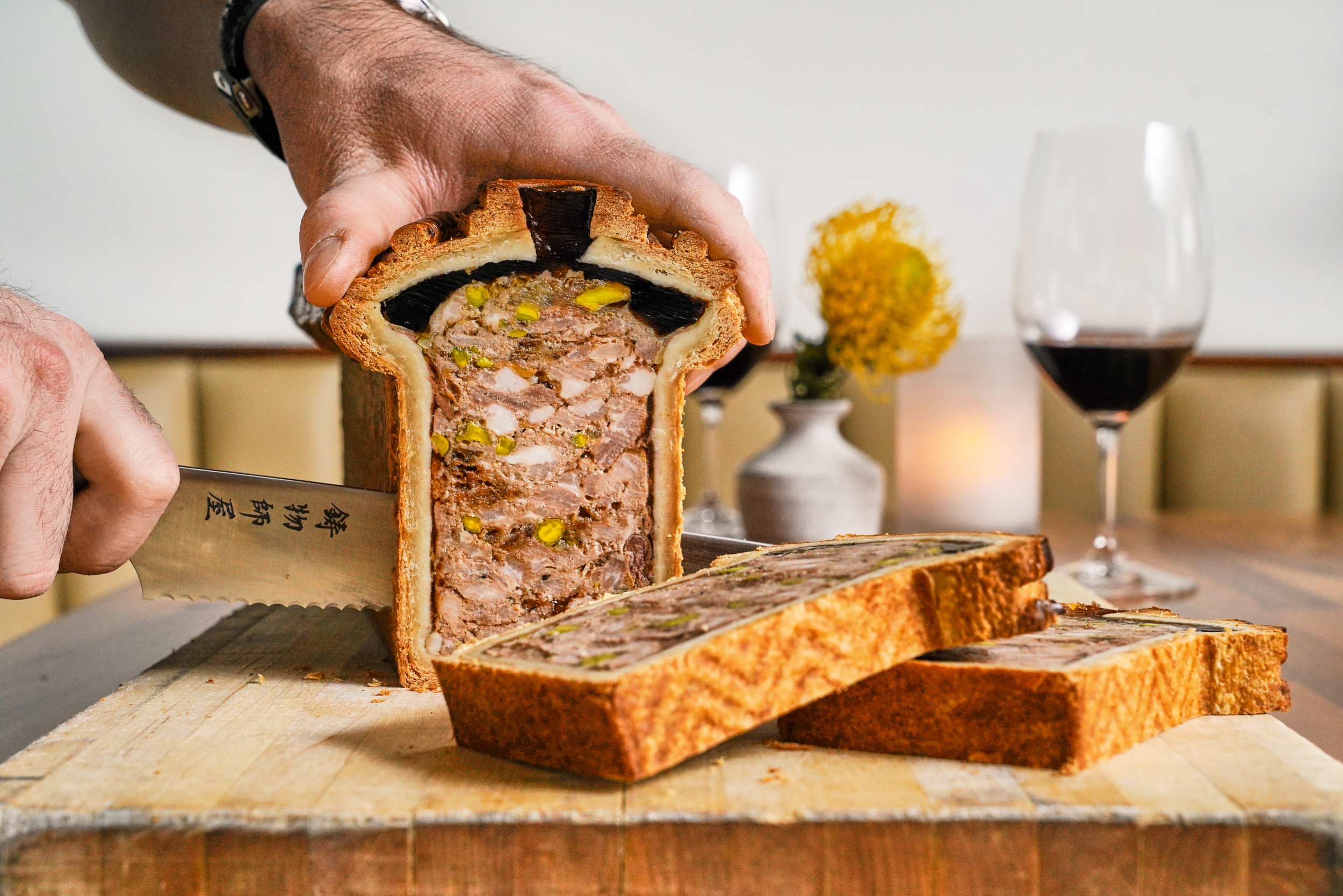 A hand is slicing a loaf of meat pie with a knife on a wooden board. The pie has a detailed filling, and two glasses of red wine are on the table nearby.