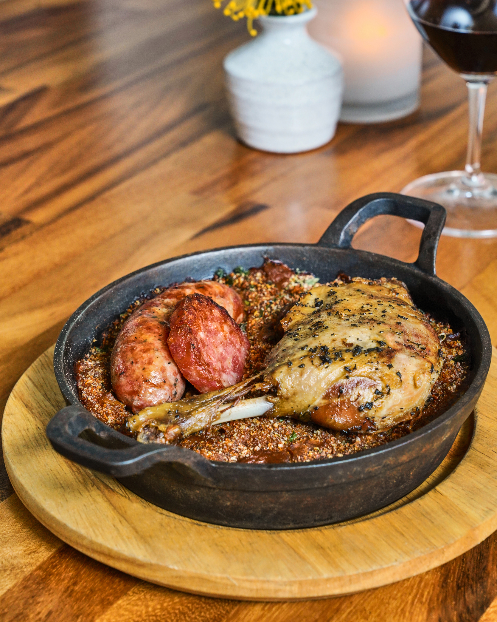 The image shows a cast iron skillet with a baked dish containing a sausage and a seasoned meat piece, likely duck, on a wooden table with a glass of red wine nearby.