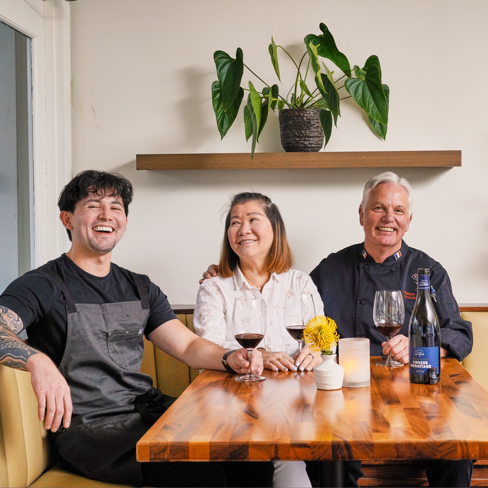 Three people sit at a wooden table with wine glasses, smiling. A plant is on a shelf behind them, and there are flowers on the table. The setting is cozy and bright.