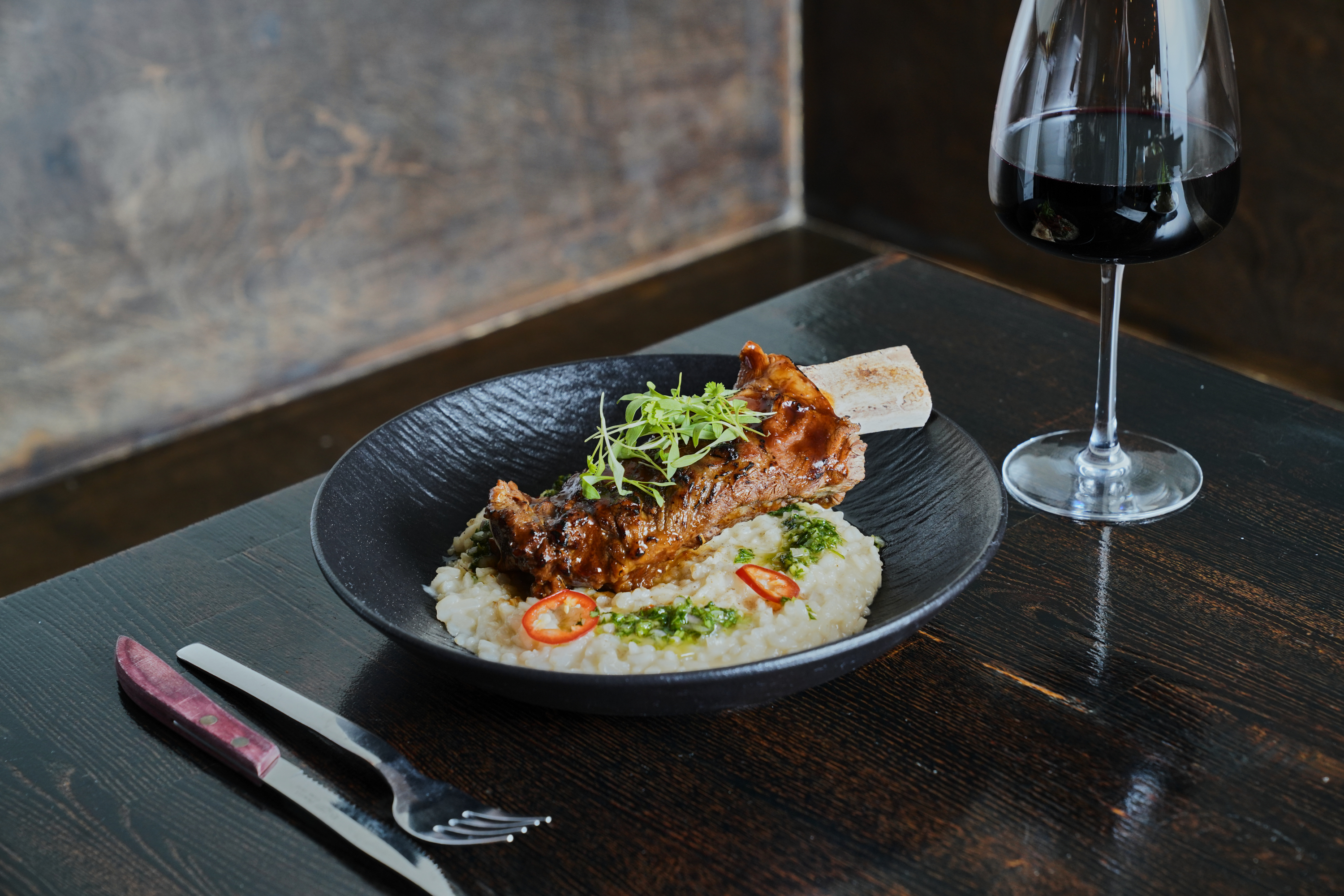 A black bowl with braised meat on risotto, garnished with greens and sliced chili, sits on a dark wooden table next to a glass of red wine.