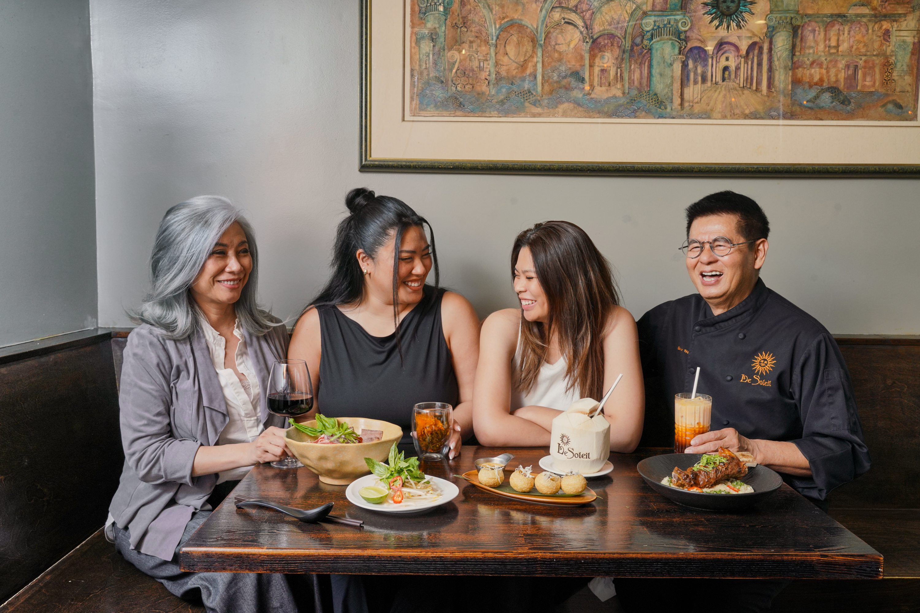 A group of four people sit at a table smiling, with dishes and drinks in front of them, including a bowl of soup, a plate with appetizers, and beverages.