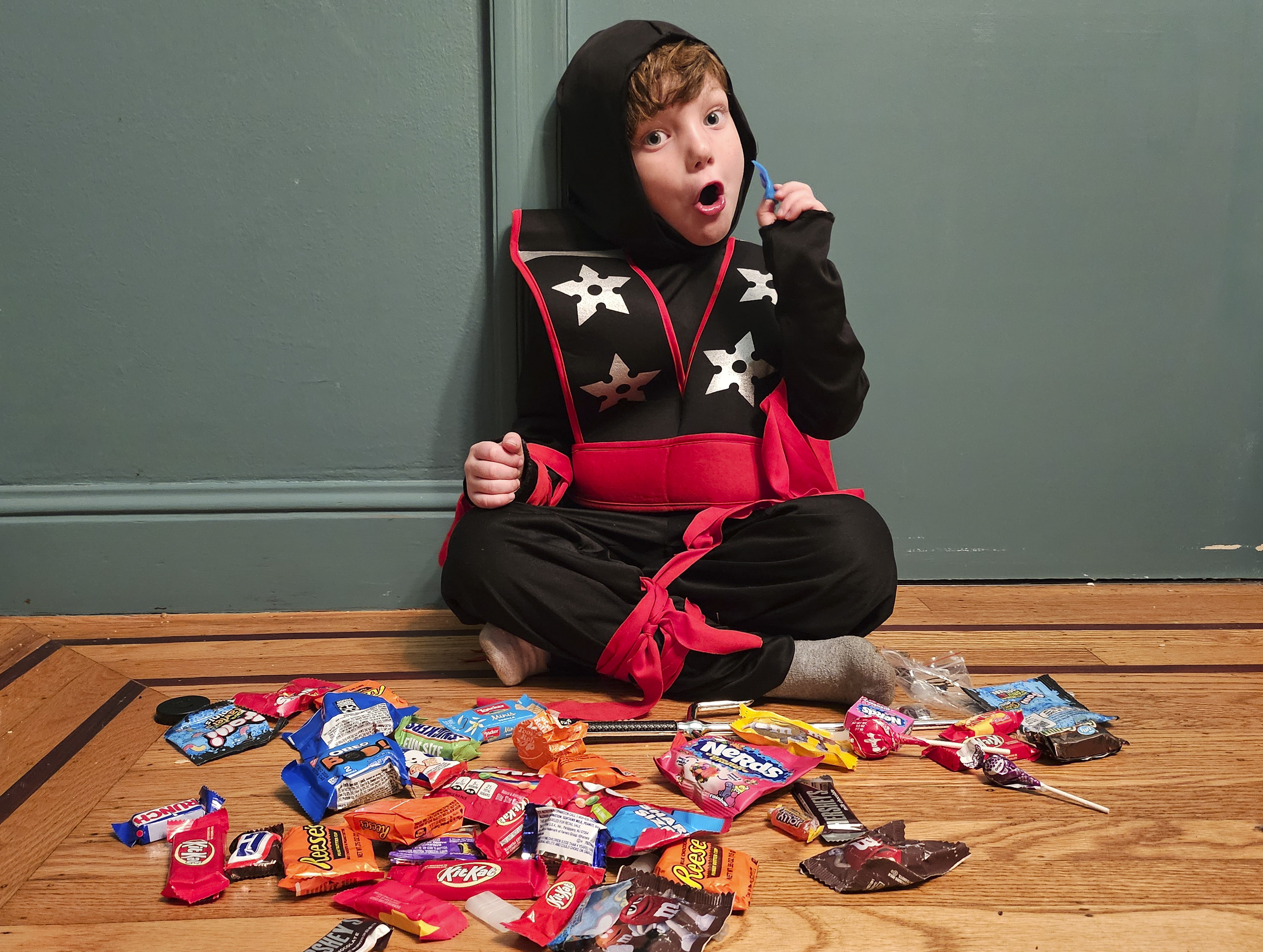 A kid in a ninja costume (black with a hood, silver stars on the chest, and a red belt) sits on the floor with a multicolor array of candy on the ground in front of him.