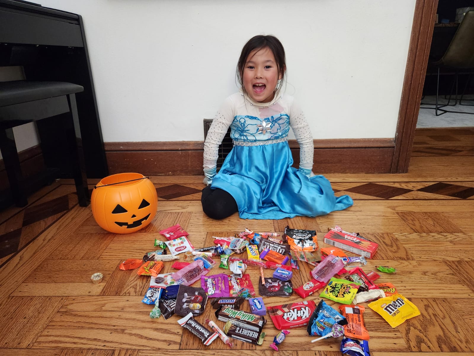 A kid in a blue dress with white long sleeves sits on the floor with multicolored candy in front of her and an orange jack-o-lantern candy bucket next to her.