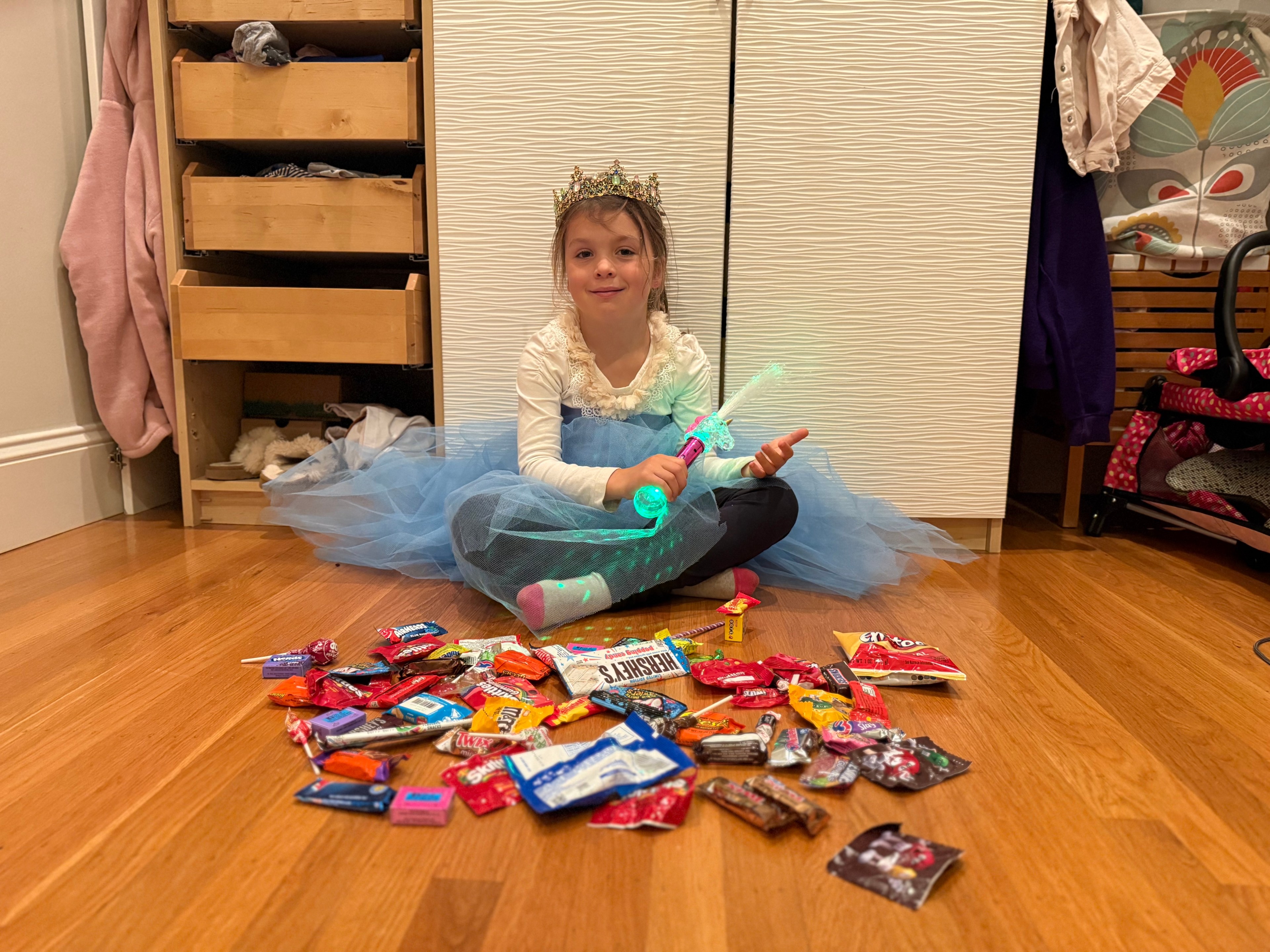 A kid in a blue dress with white long sleeves sits on the floor with multicolored candy in front of her.