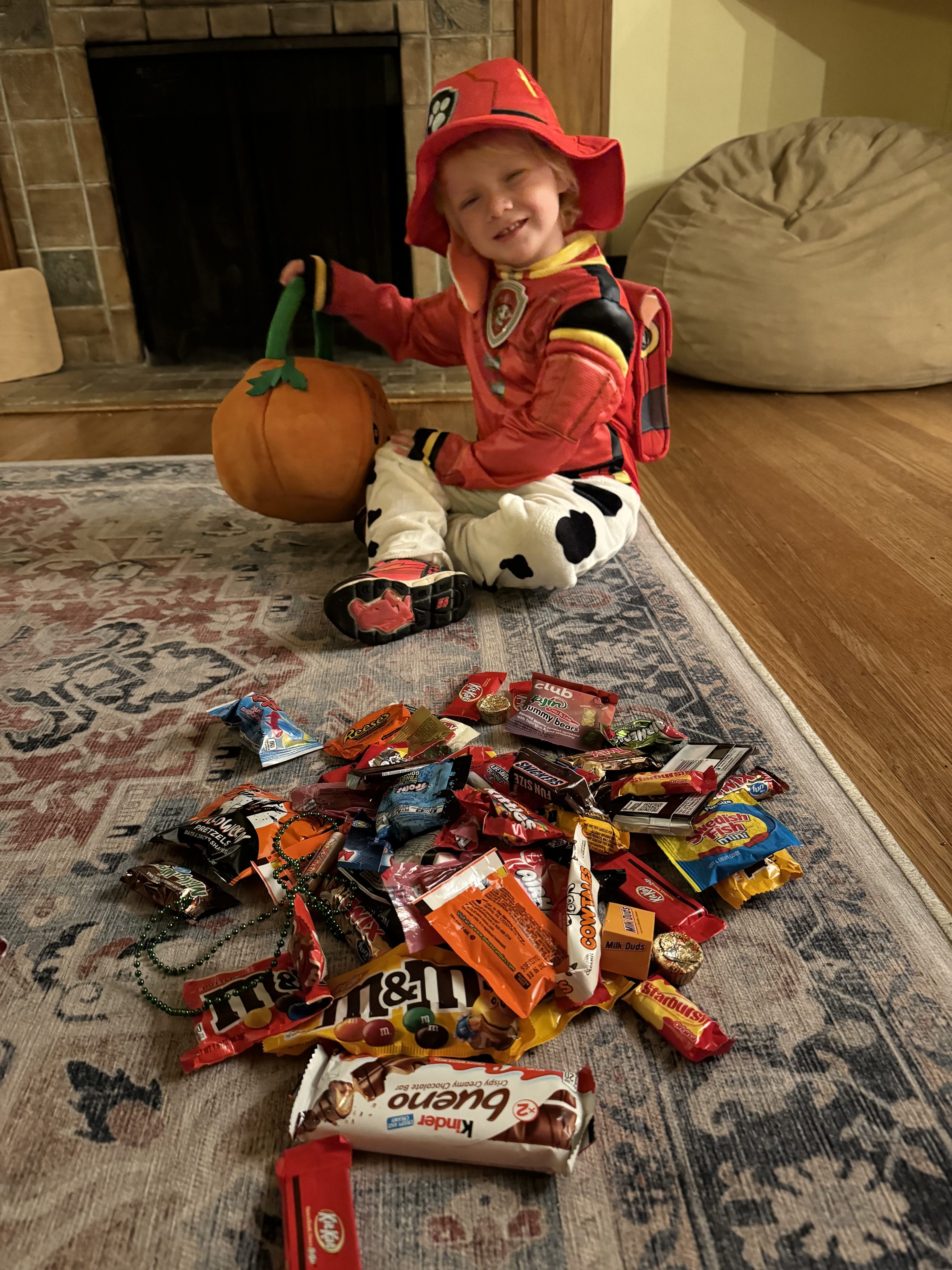 A kid in a red firefighter-esque shirt and hat and white, spotted pants sits on the ground and smiles while holding an orange Jack-o-lantern candy bucket. A multicolored spread of candy is on the floor in front of him.