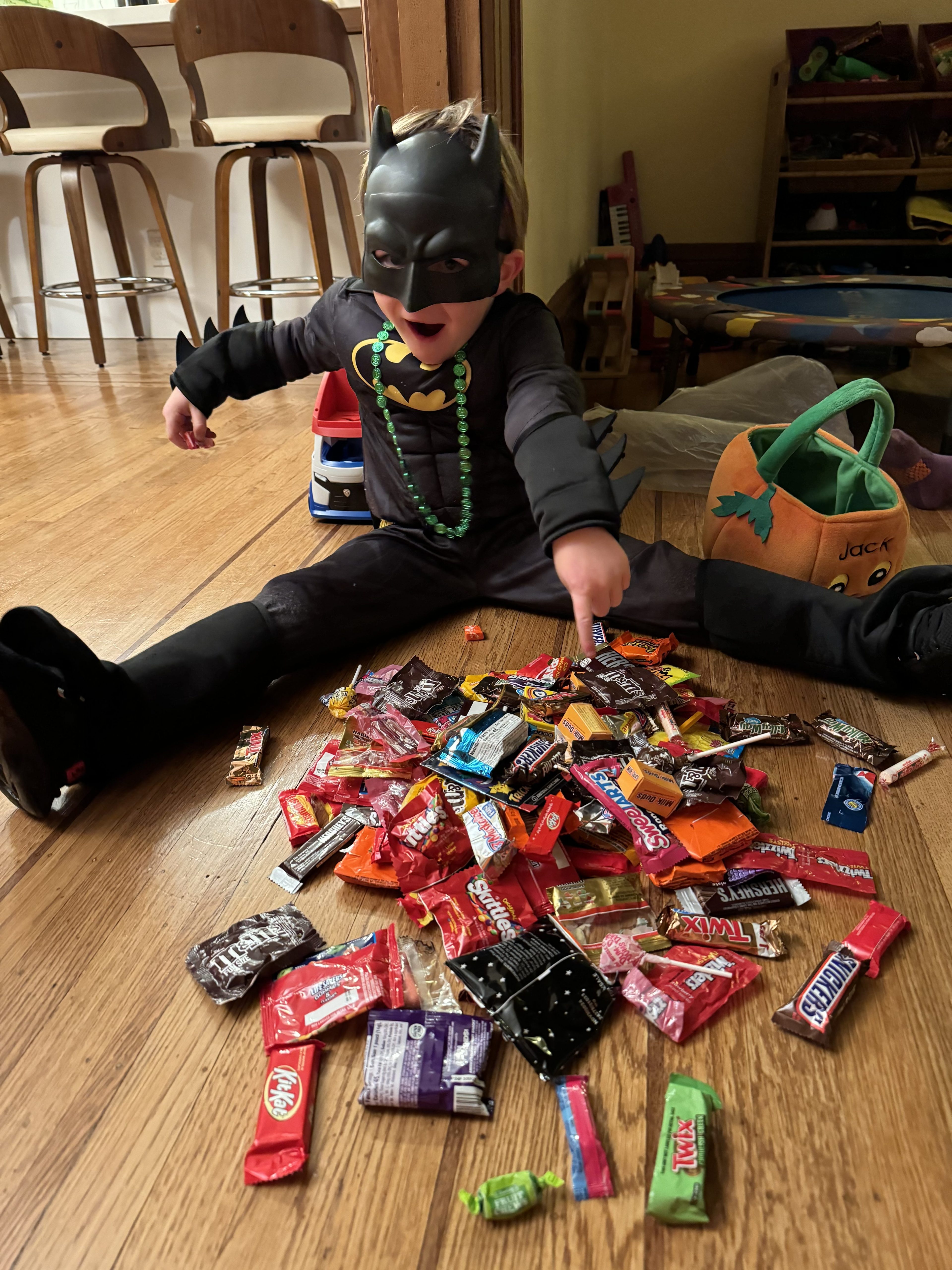 A kid in a black Batman costume and a mask sits on the ground, pointing to the spread of multicolored candy in front of him.