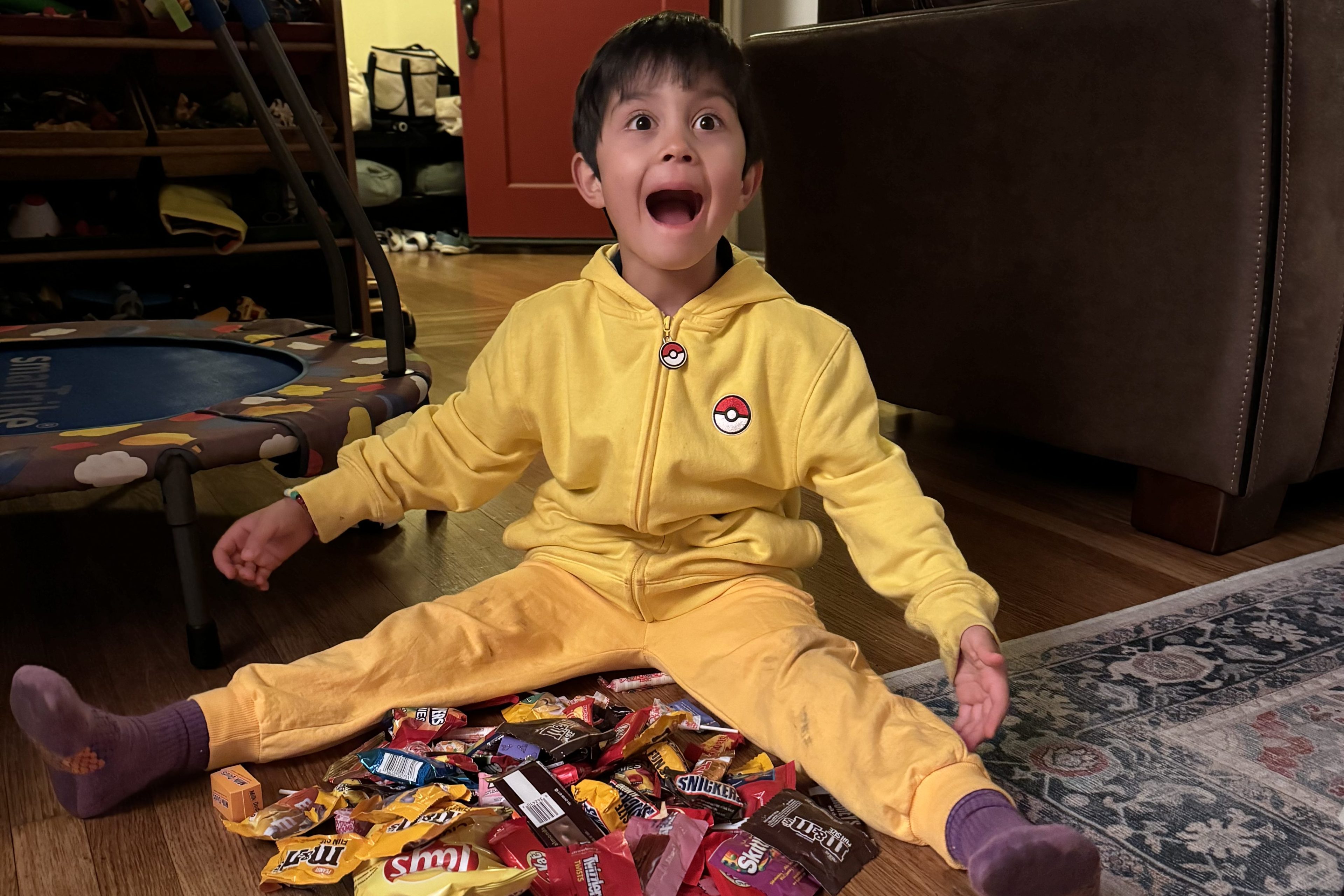 A kid with brown hair wearing a yellow zip-up sweater and yellow pants sits on the ground with candy out in front of him.