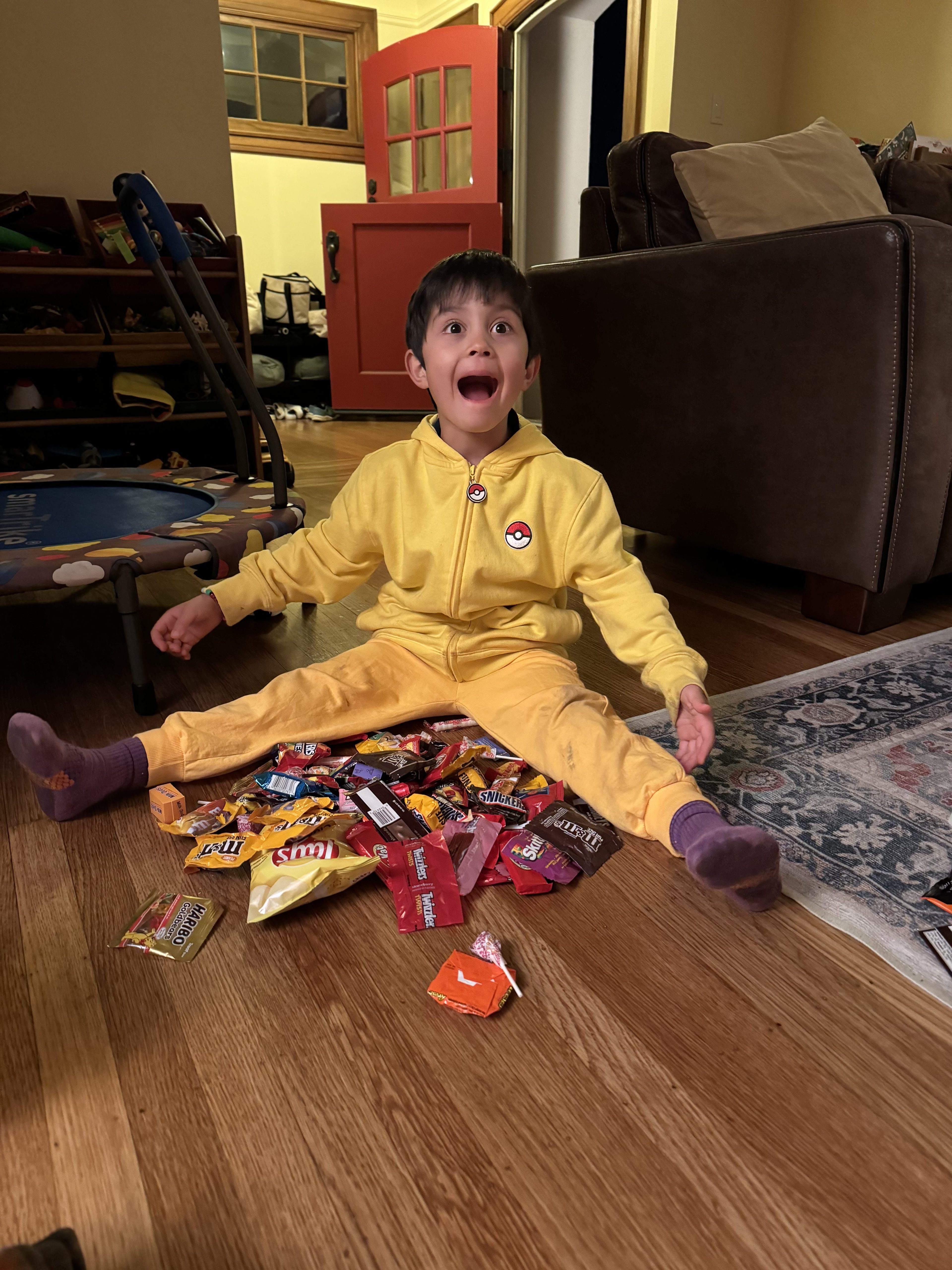 A kid with brown hair wearing a yellow zip-up sweater and yellow pants sits on the ground with candy out in front of him.