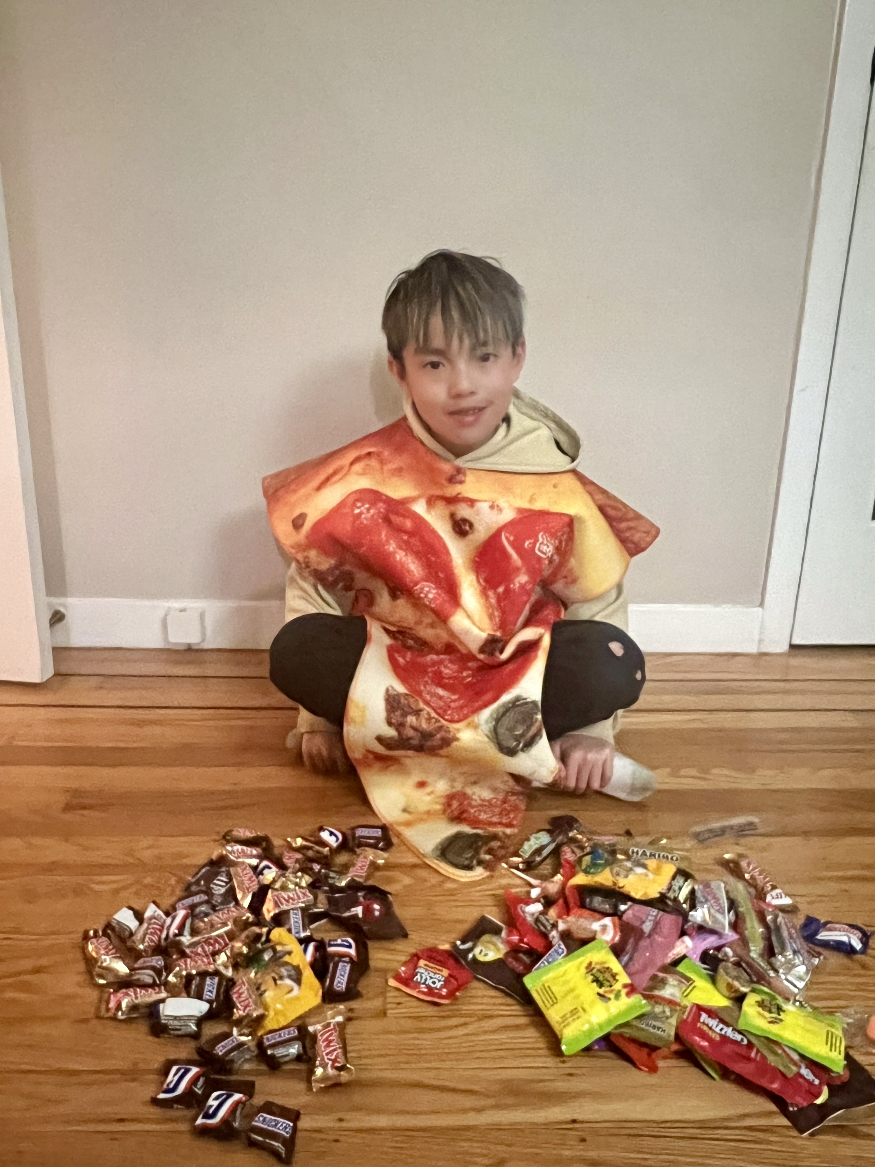 A kid dressed in a pizza costume sits on the ground against a wall with a spread of multicolored candy in front of him.