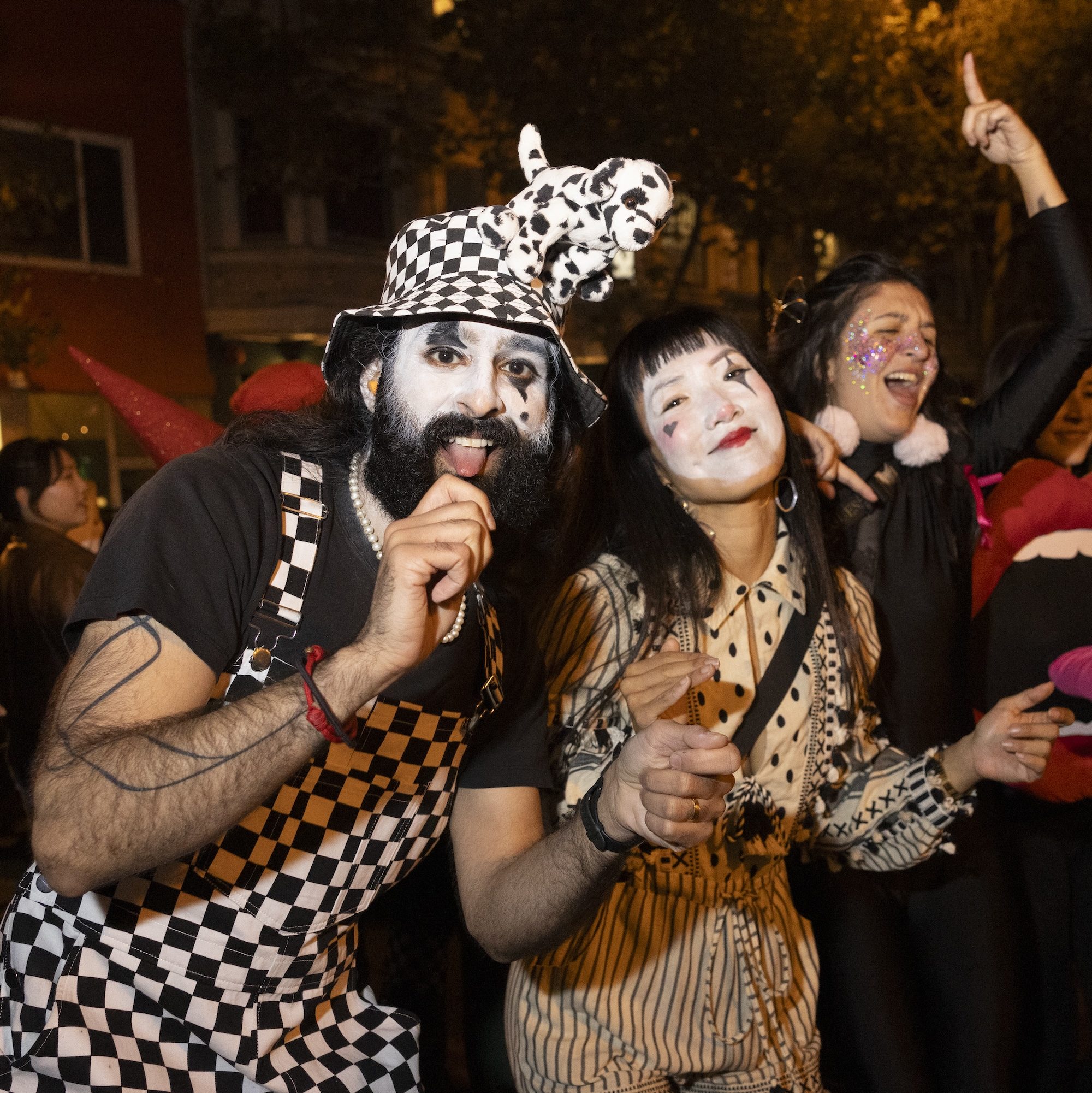 Three people in costumes and face paint are celebrating at night. One is in checkered attire, another in a striped blouse, and the third with glitter and a playful prop.