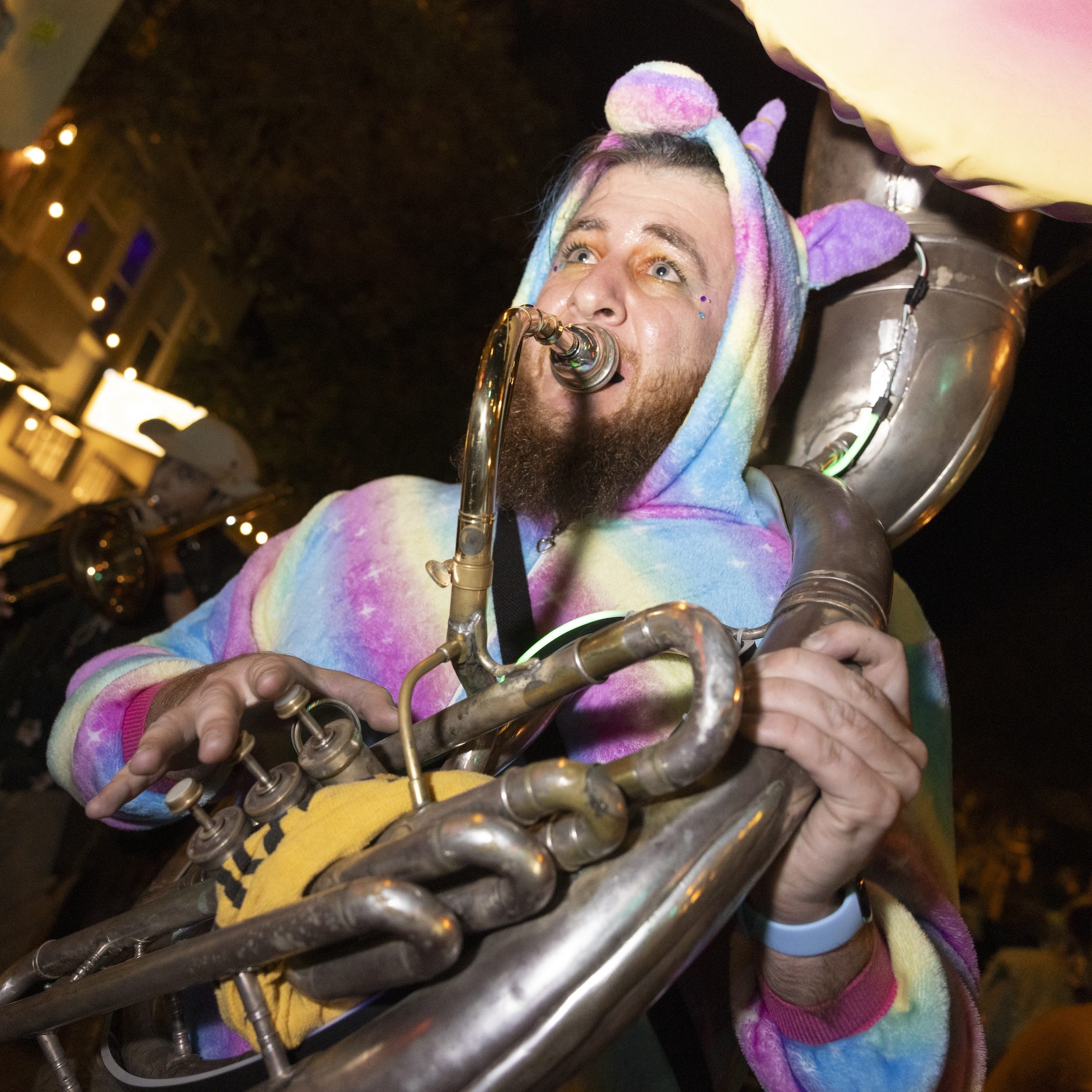 A person in a colorful rainbow unicorn onesie is playing a sousaphone at night, with twinkling lights in the background creating a festive atmosphere.