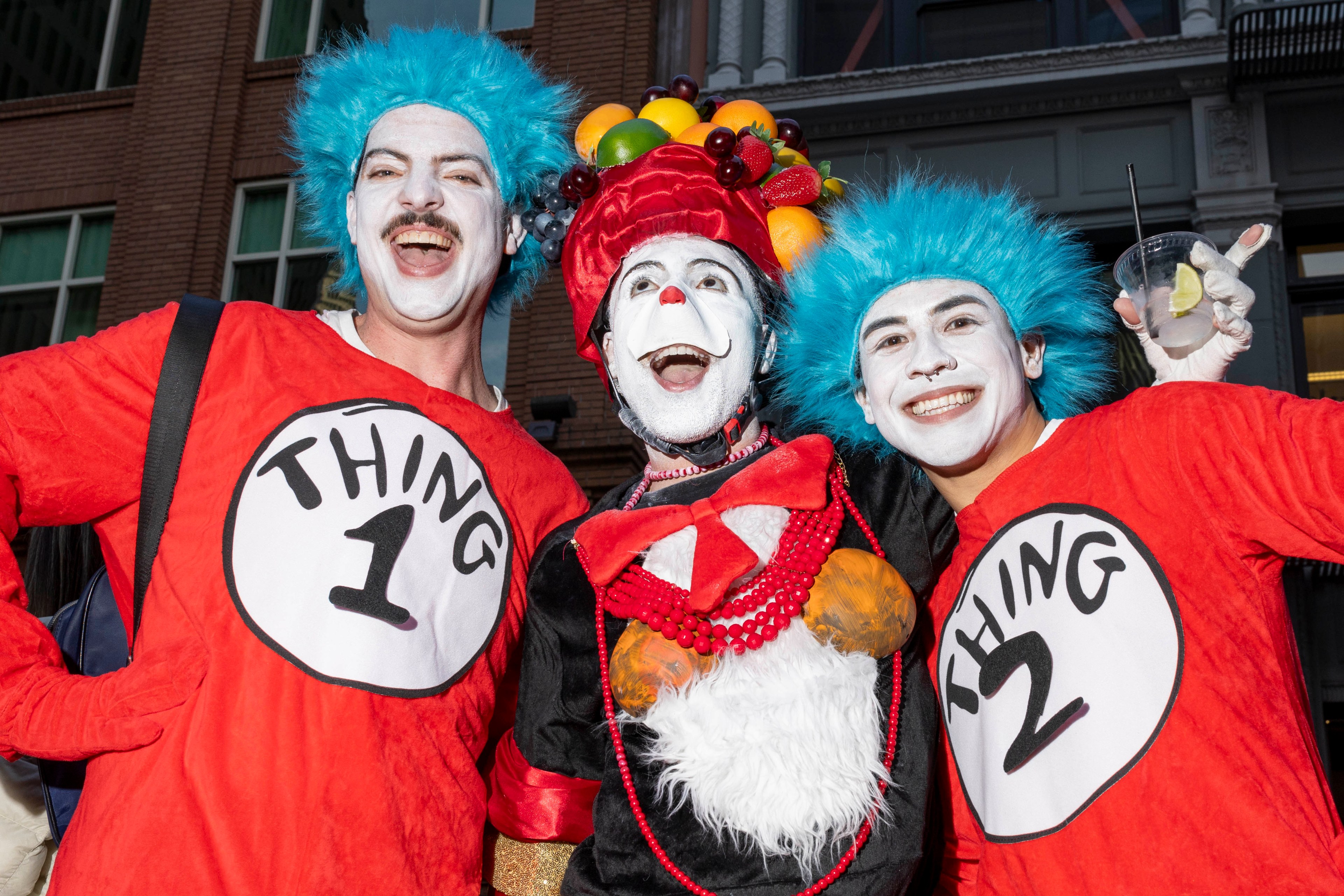 Three people in colorful costumes are smiling; two dressed as &quot;Thing 1&quot; and &quot;Thing 2&quot; with blue wigs, and one in a fruit-adorned hat and red outfit.