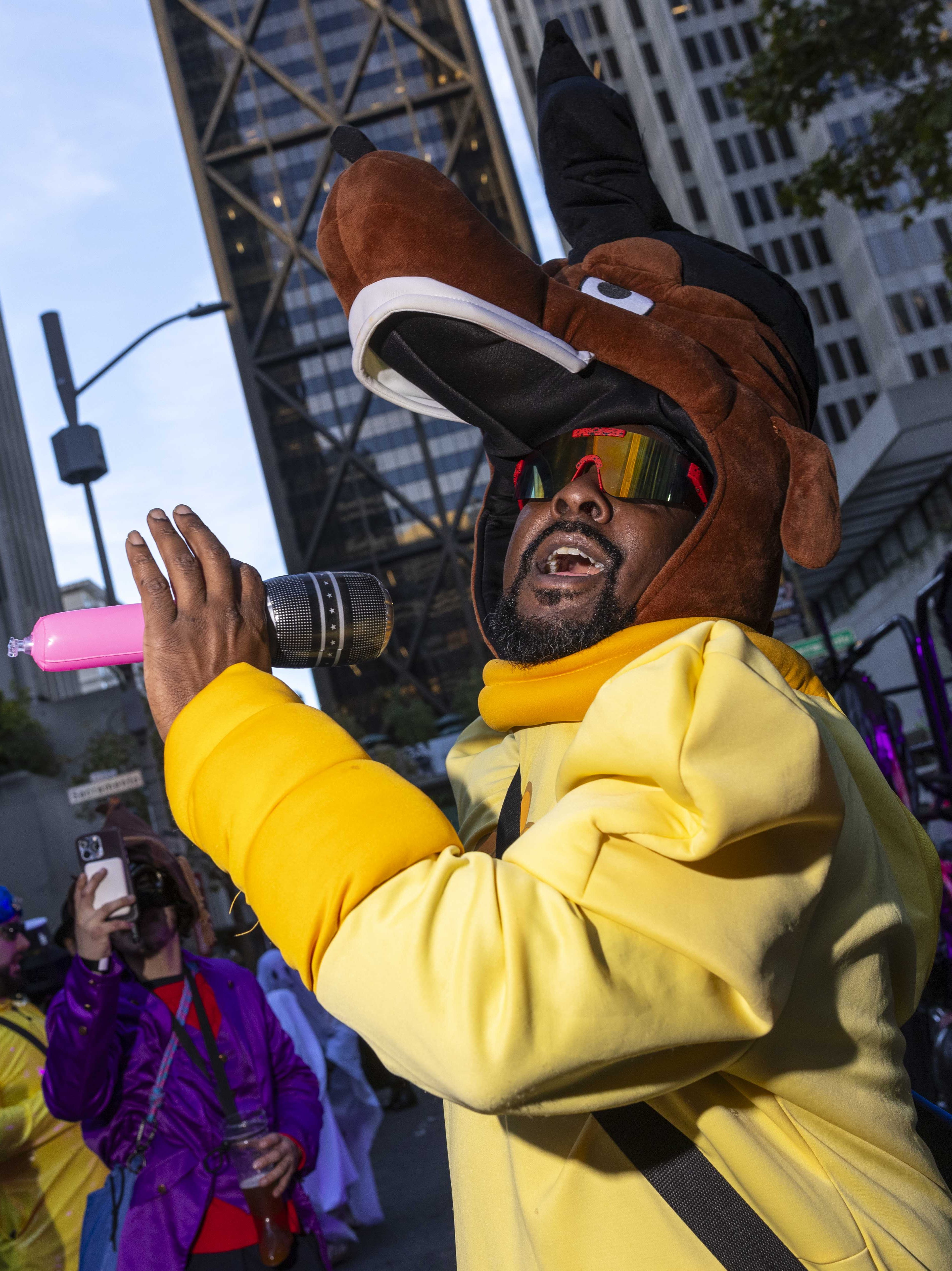 A person in a Scooby-Doo costume sings into a microphone, surrounded by people in colorful costumes on a busy city street with tall buildings.