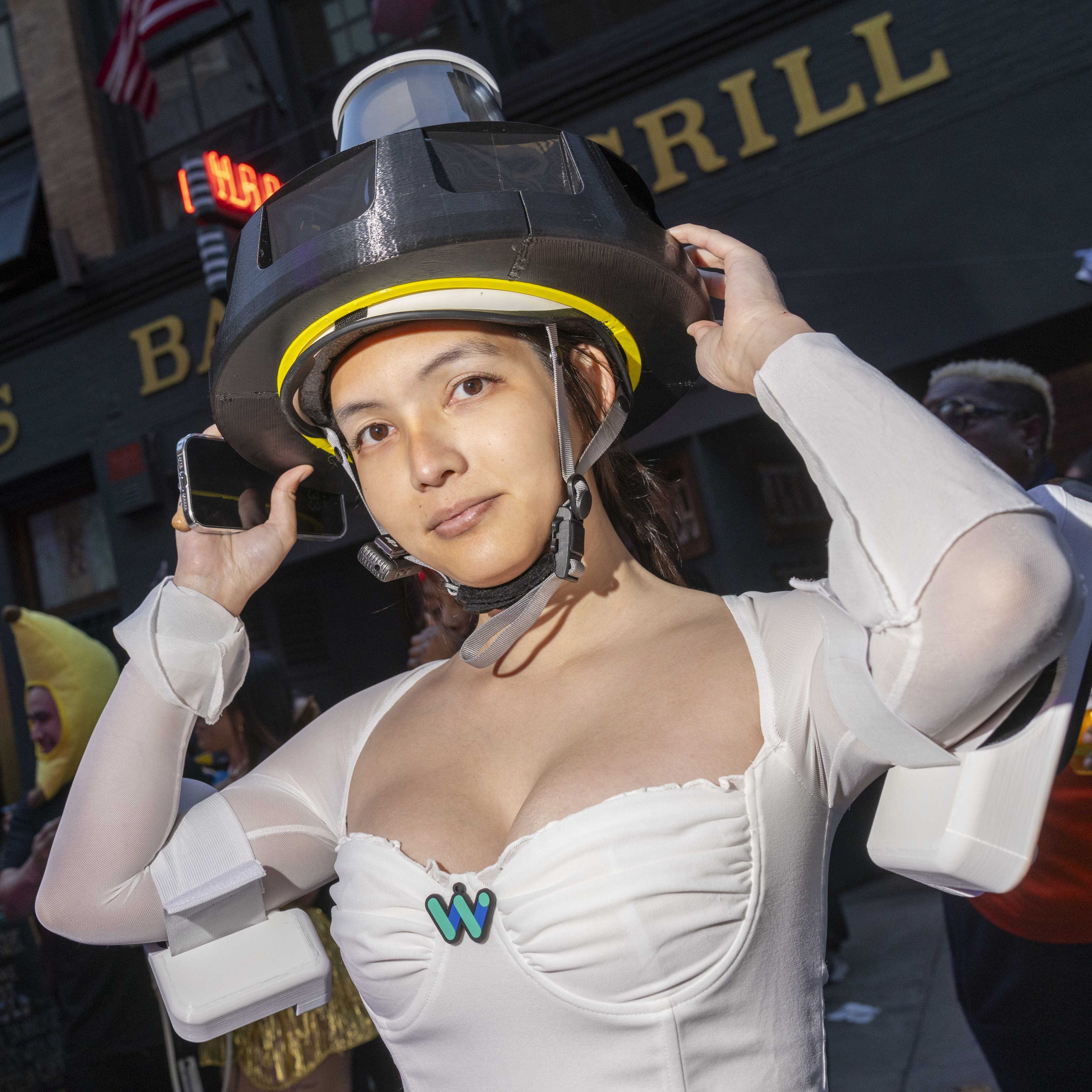 A person wearing a creative white costume with a large helmet stands outdoors. Behind them are people in various costumes and a building labeled &quot;BAR &amp; GRILL.&quot;