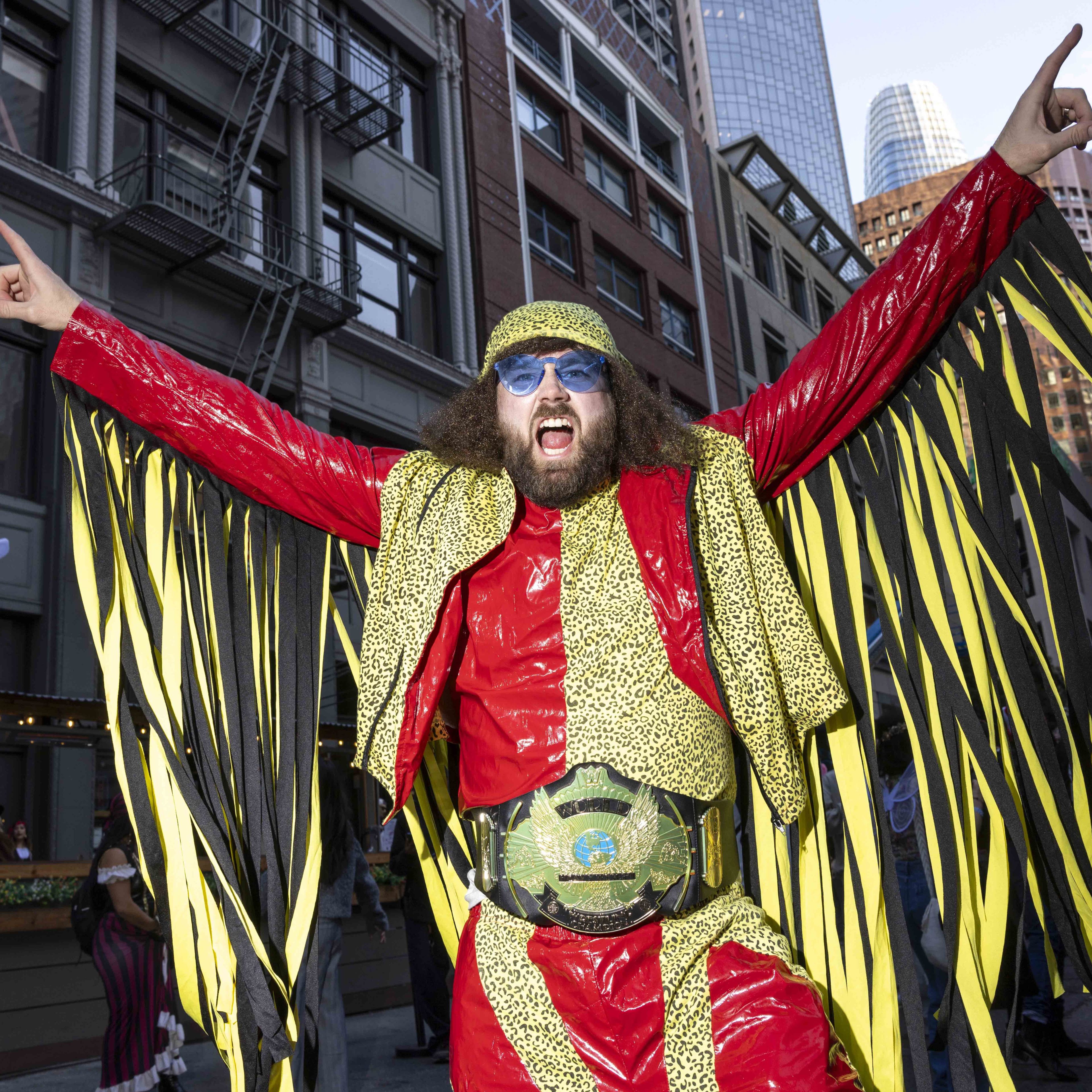 The image shows a person excitedly posing in a flamboyant outfit with red and leopard print patterns, blue sunglasses, and a large championship belt. Buildings are visible behind them.