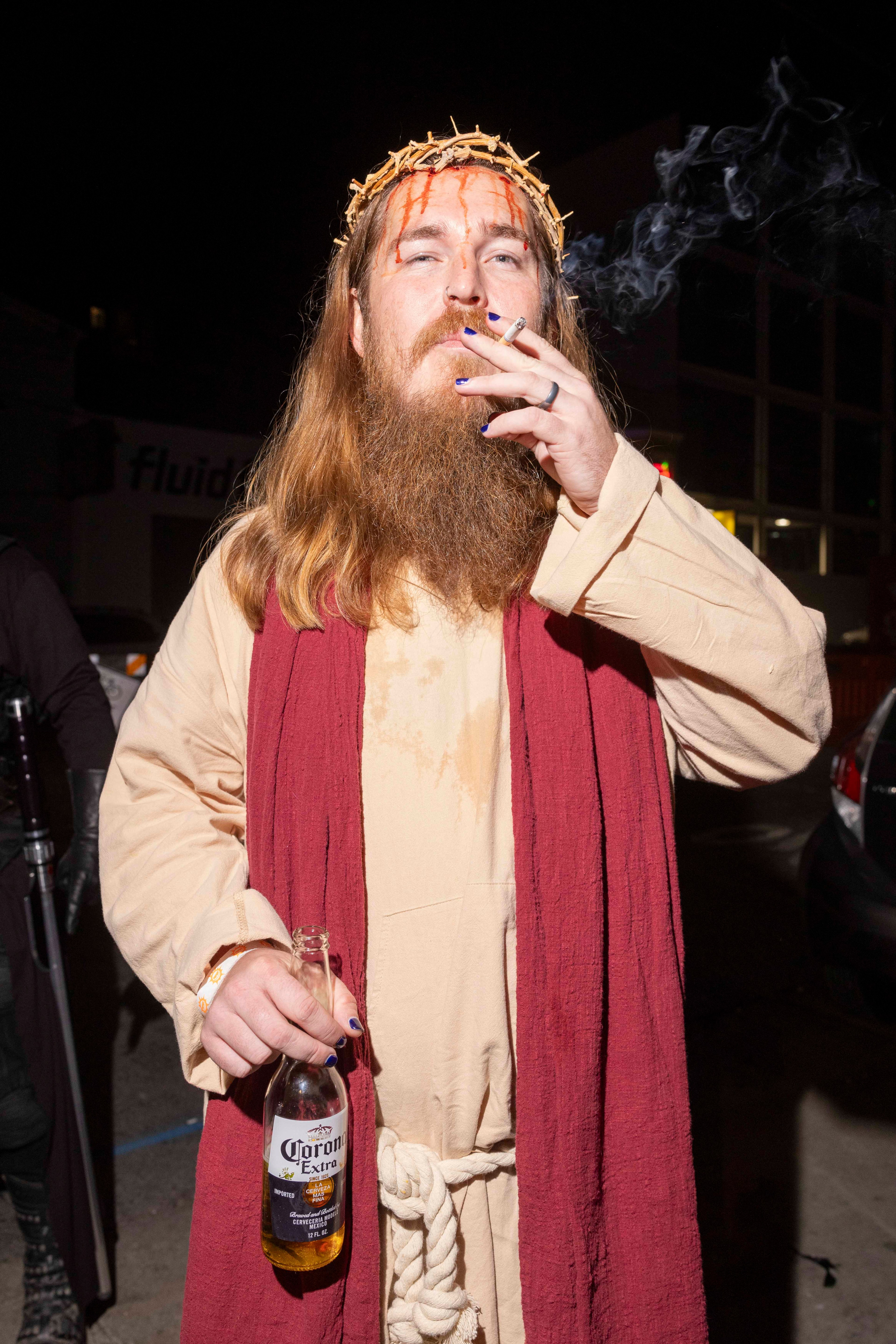 A person dressed as Jesus smokes a cigarette, holds a beer, and wears a crown of thorns. They have long hair, a beard, and painted blue nails.