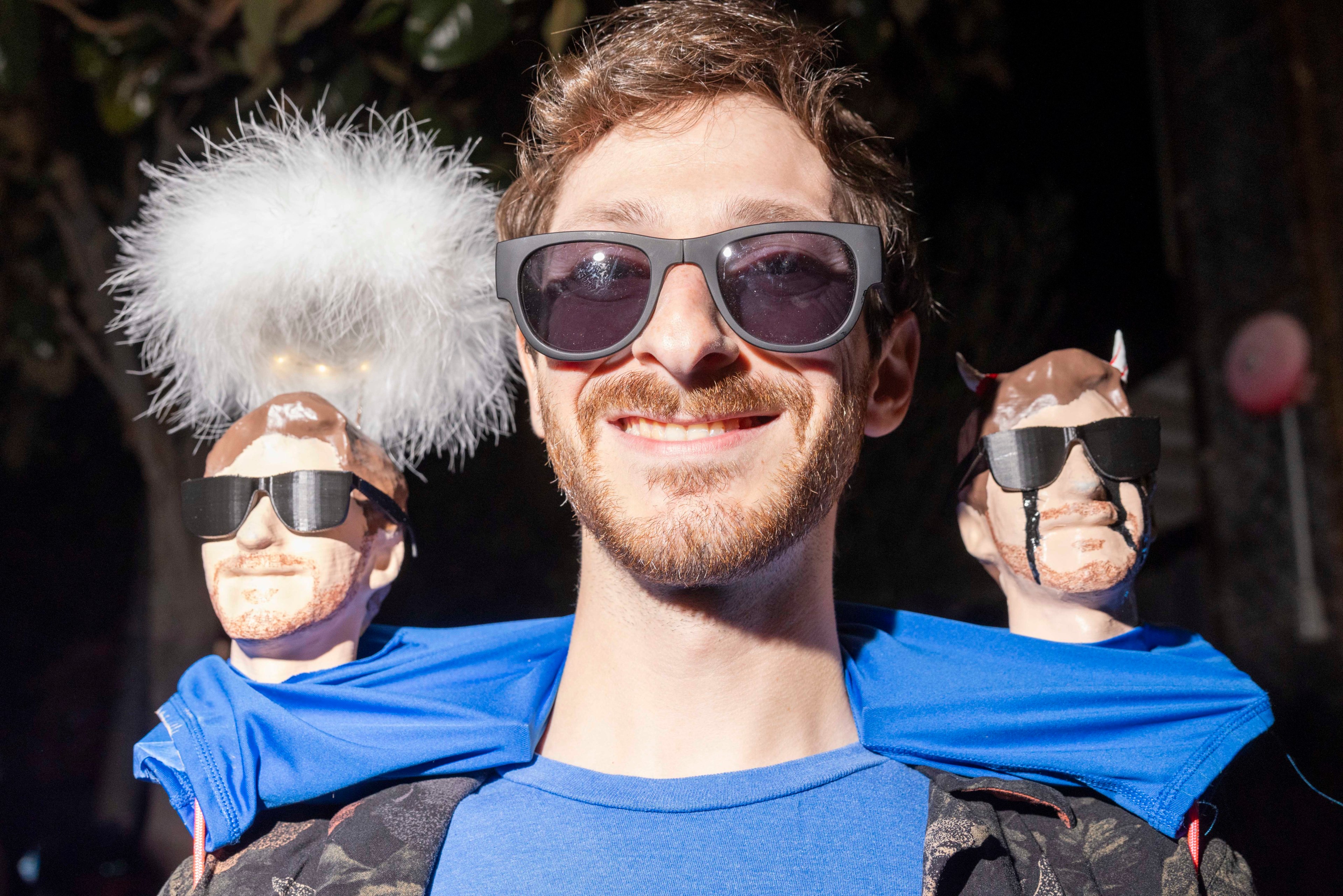 A man wearing sunglasses smiles, flanked by two small models of himself. One model has angelic features, with a fluffy halo, and the other has devil horns.