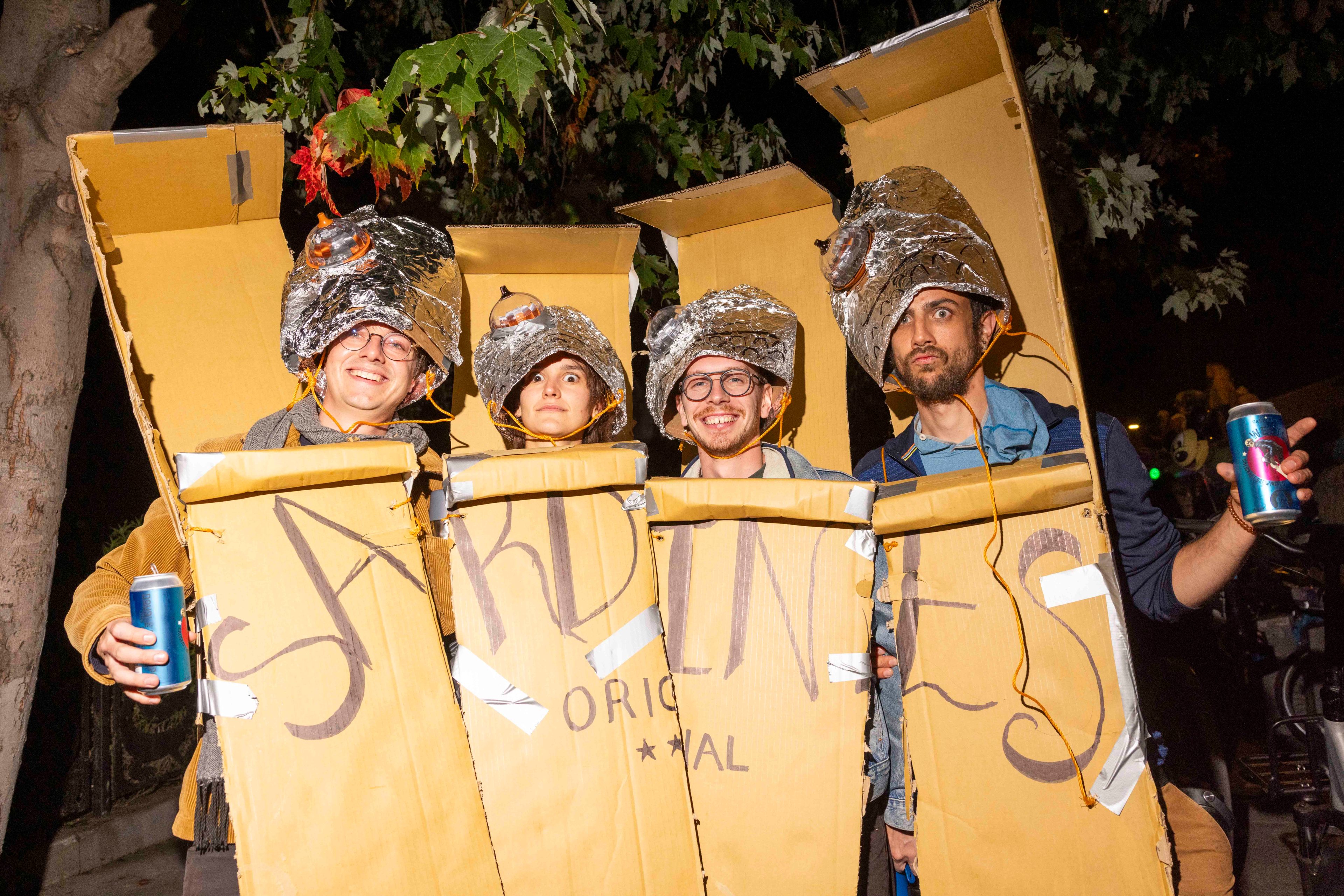 Four people, standing together, wear cardboard box costumes resembling sardine cans. They hold drinks and have foil hats, with trees in the background.