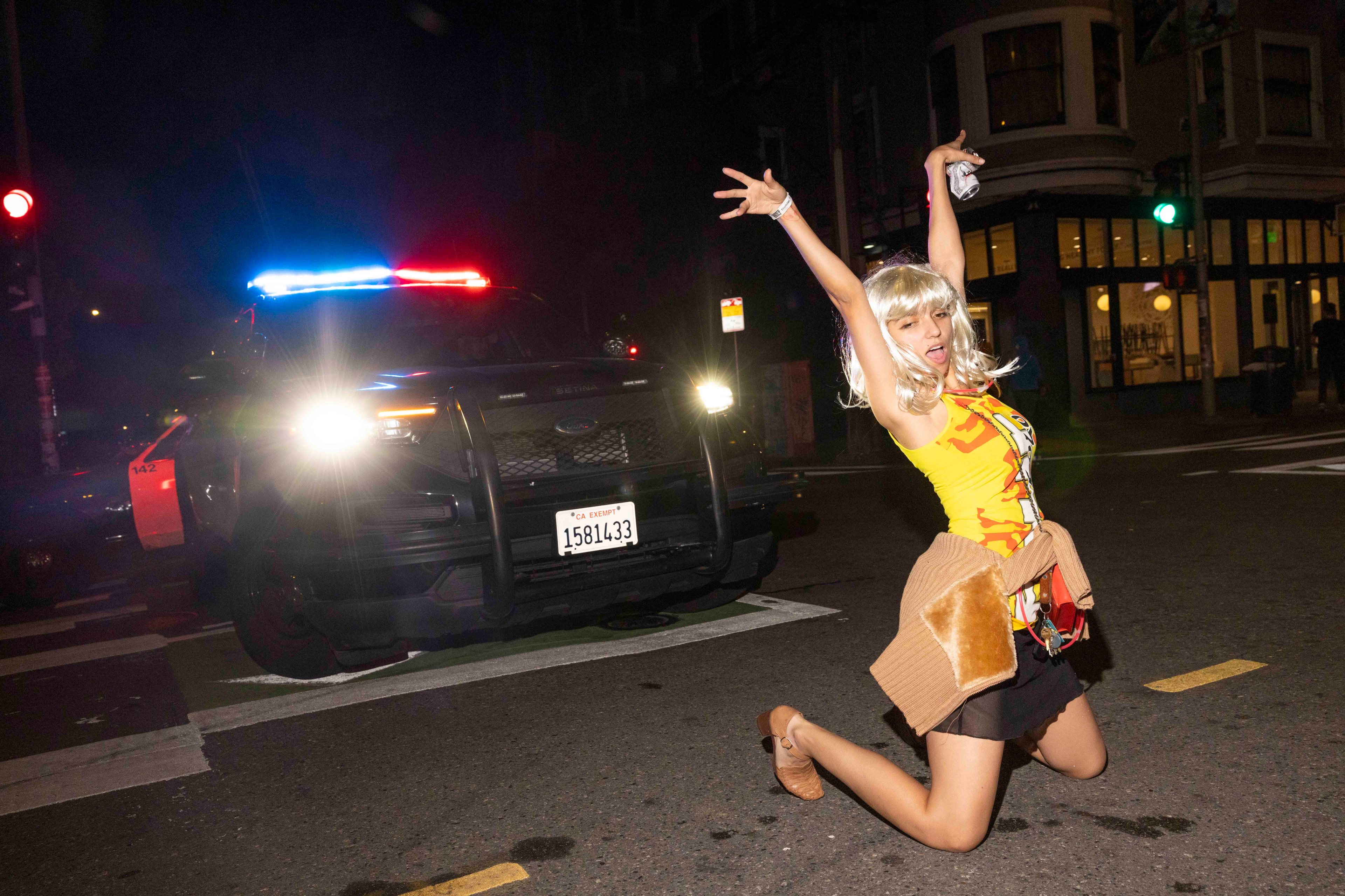 A person in a colorful outfit poses energetically on a street at night, as a police car with flashing lights is seen behind them.