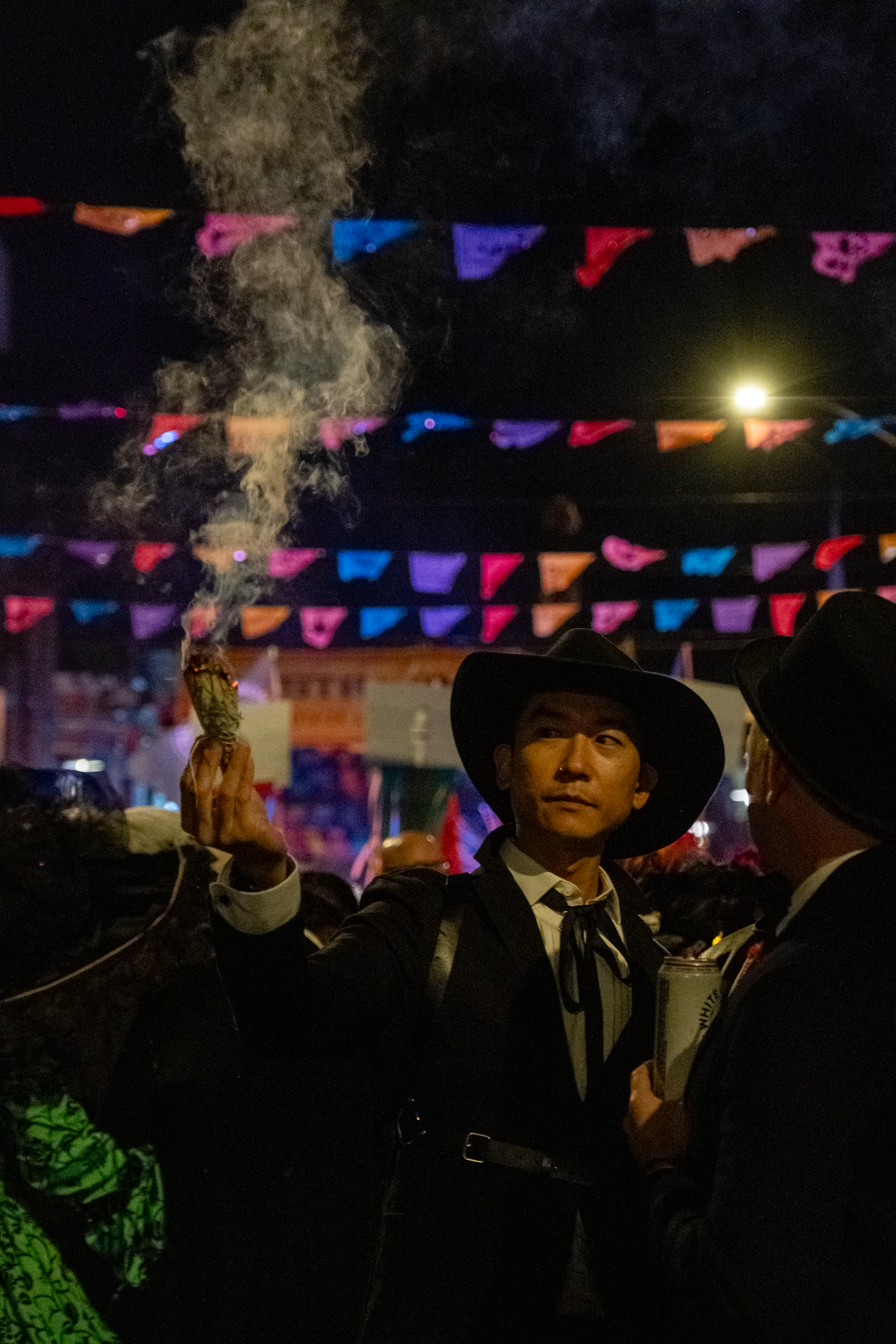 A person in a dark suit and hat holds a smudge stick emitting smoke at night. Colorful banners hang above them, illuminated by a streetlamp.